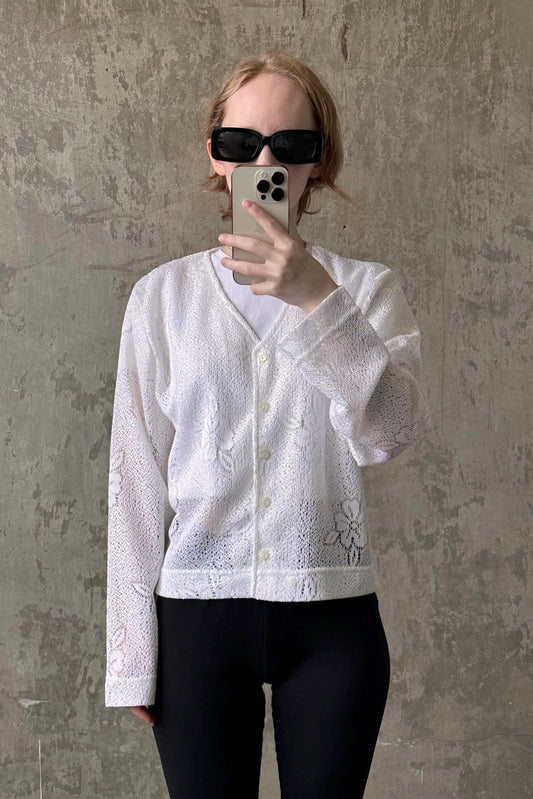 A person wearing a white lace cardigan and sunglasses is taking a selfie with a smartphone.