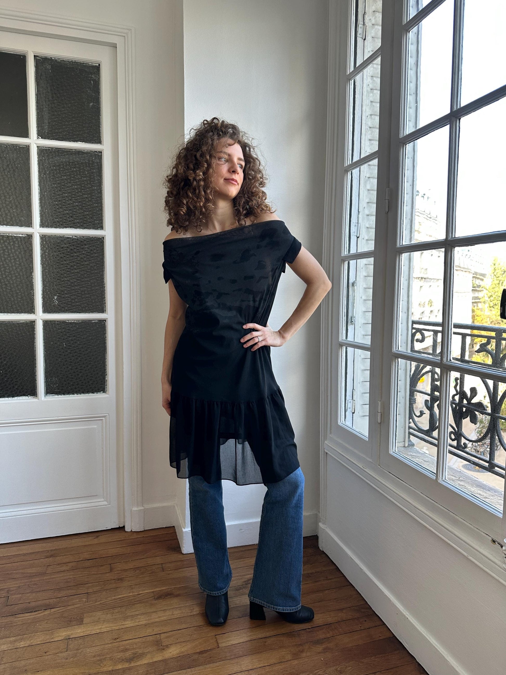 A woman with curly hair stands in a room, wearing a black off-the-shoulder dress and blue jeans.