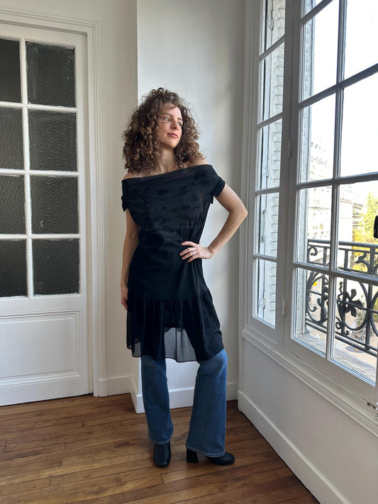 A woman with curly hair stands in a room, wearing a black off-the-shoulder dress and blue jeans.