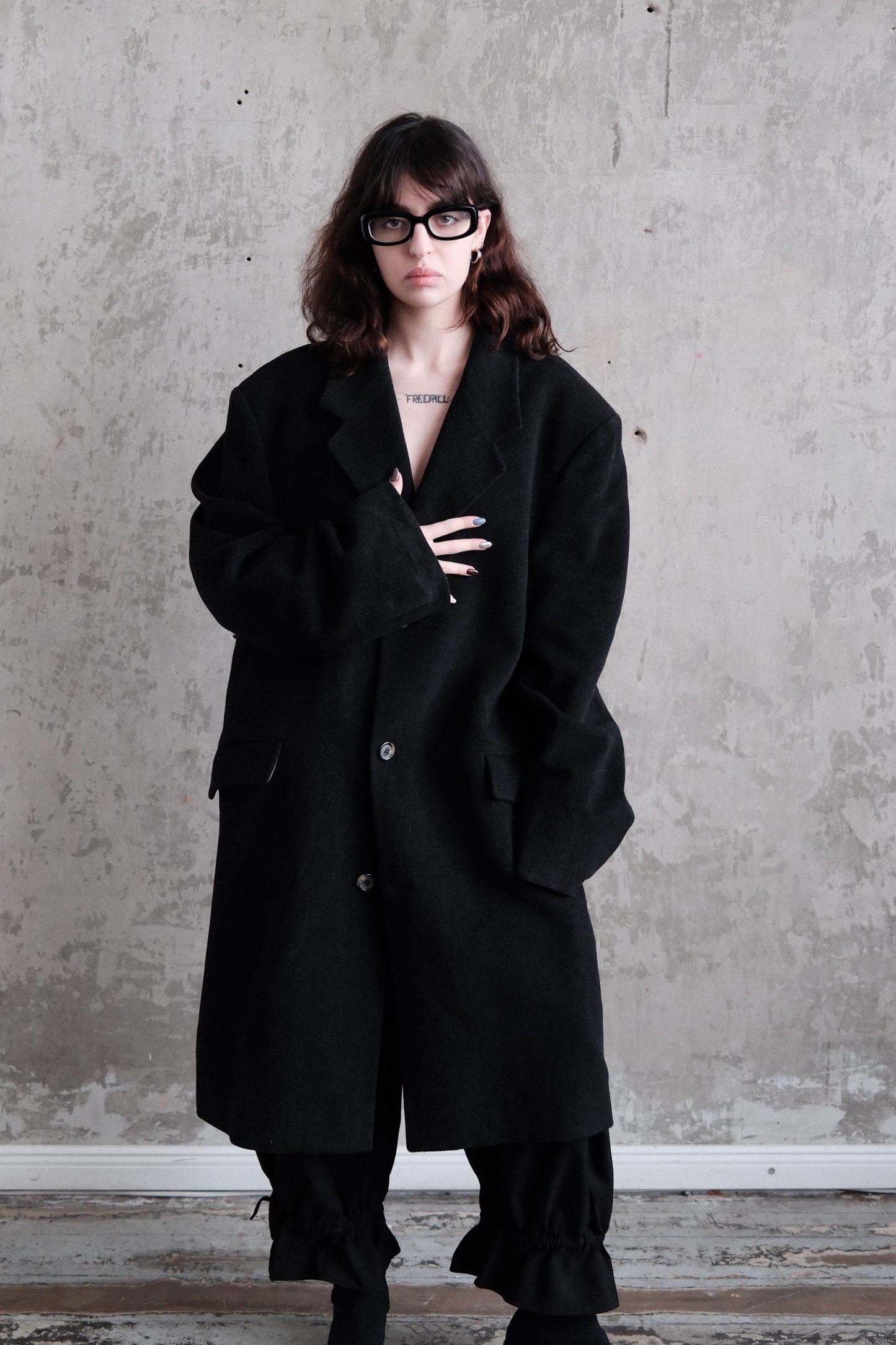 A woman wearing a black coat and glasses stands against a plain gray wall, looking to the side with a serious expression.