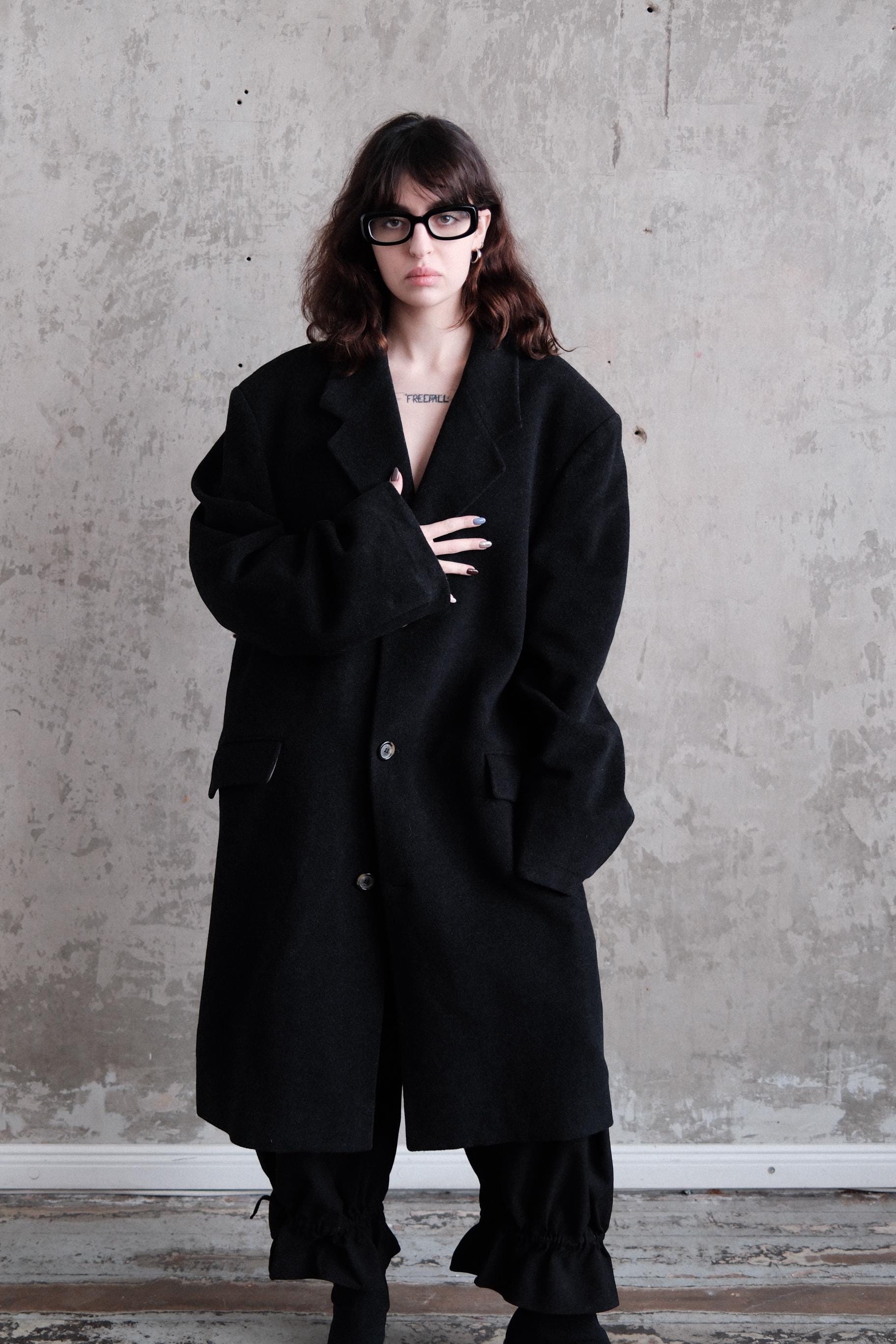 A woman wearing a black coat and glasses stands against a plain gray wall, looking to the side with a serious expression.