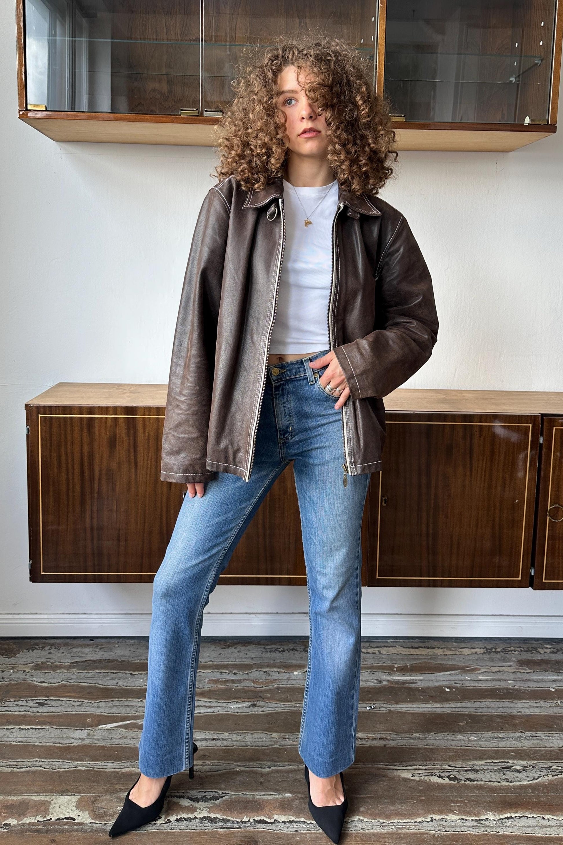 A woman with curly hair wearing a brown leather jacket, white shirt, and blue jeans stands in a room with wooden cabinets.