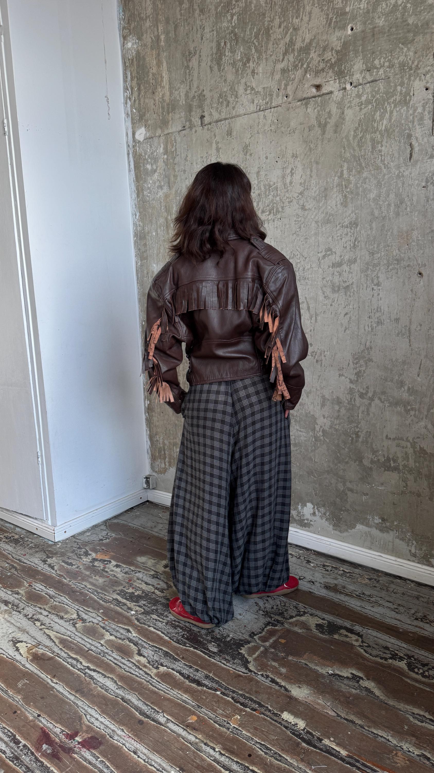 A person wearing a brown leather jacket with fringe details and plaid pants stands in a room with a concrete wall.