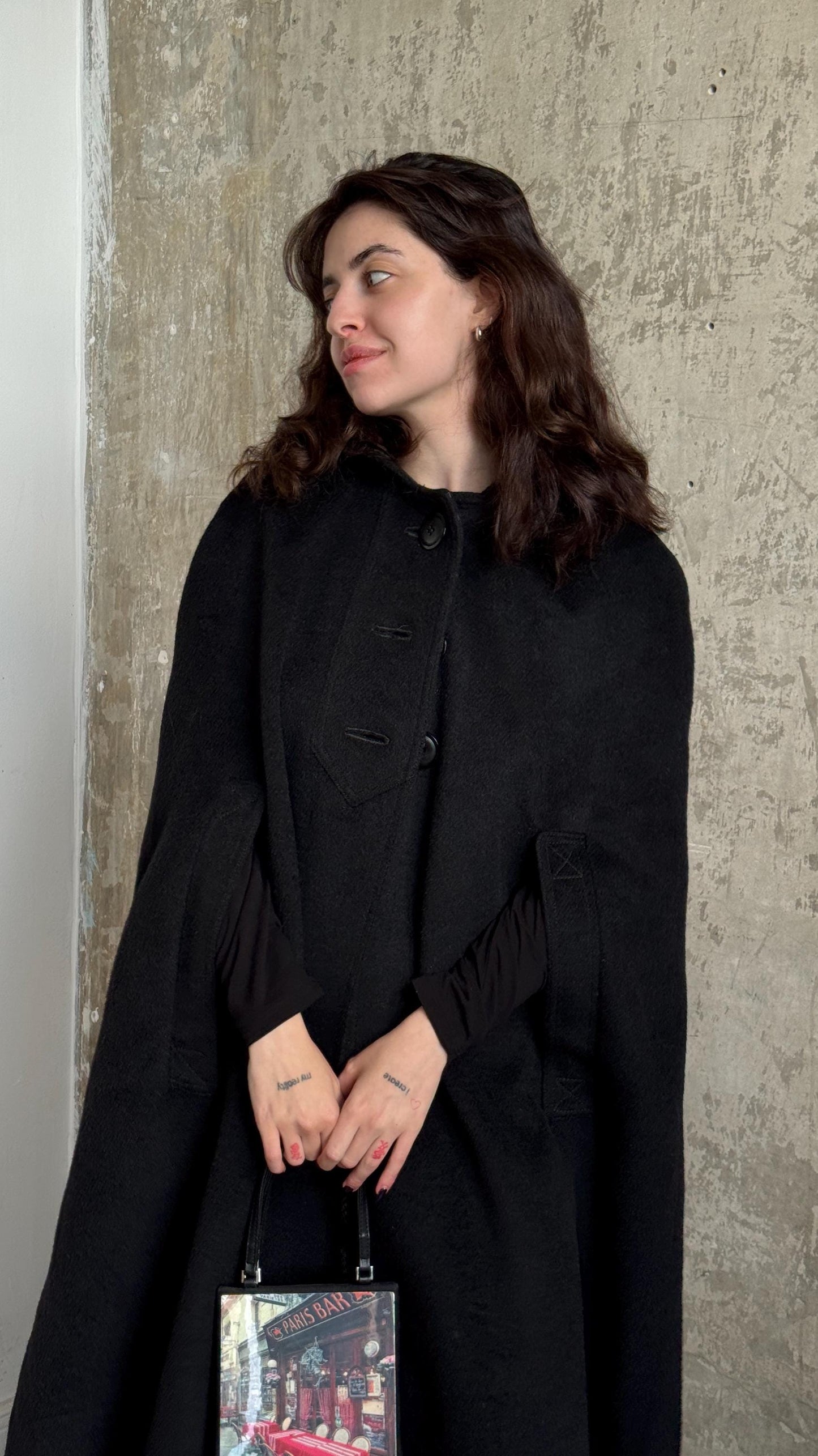 A woman in a black coat stands against a concrete wall, holding a bag with a picture on it.