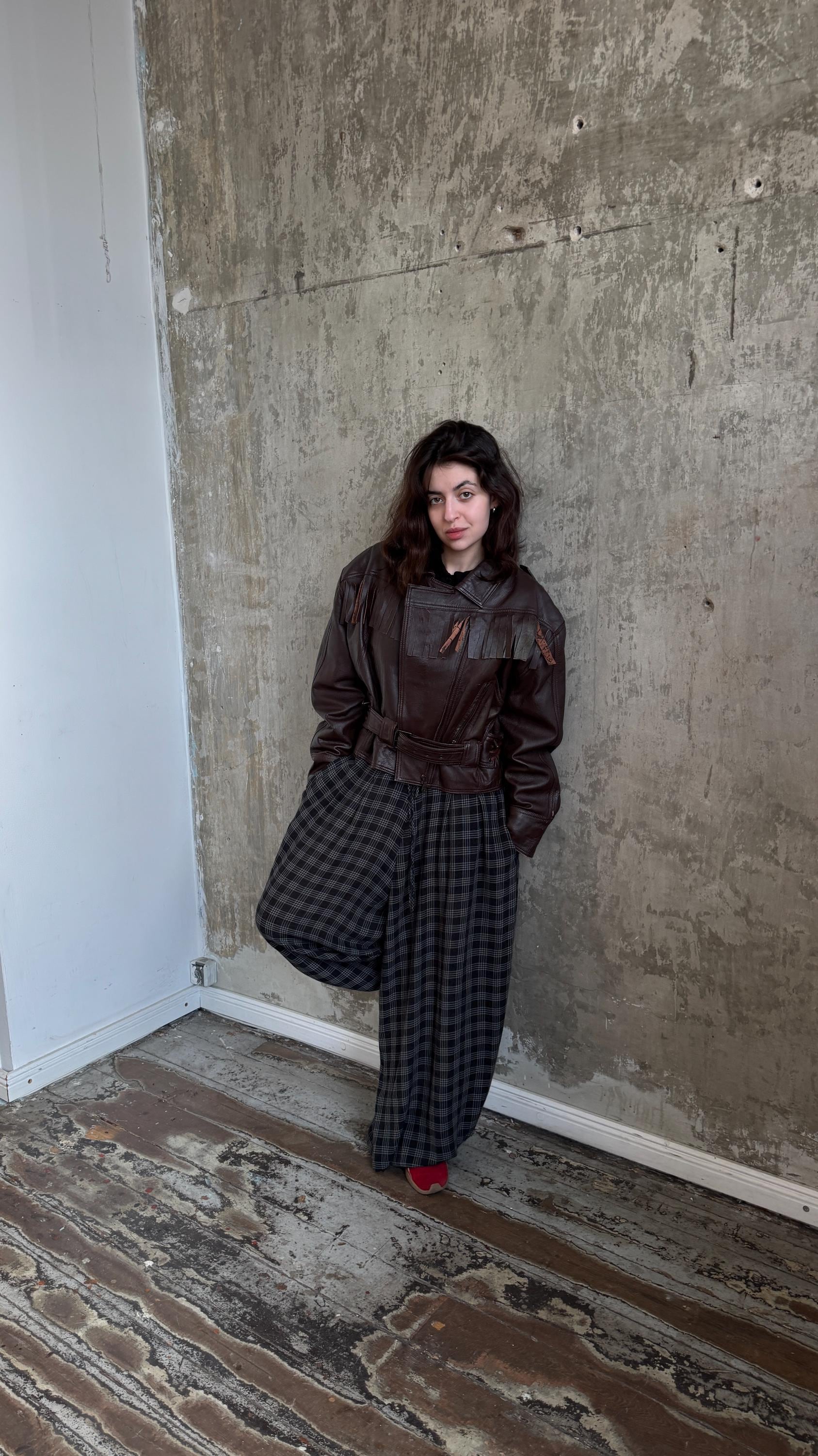 A woman in a brown leather jacket and plaid pants stands against a concrete wall, leaning against it with one leg in the air.