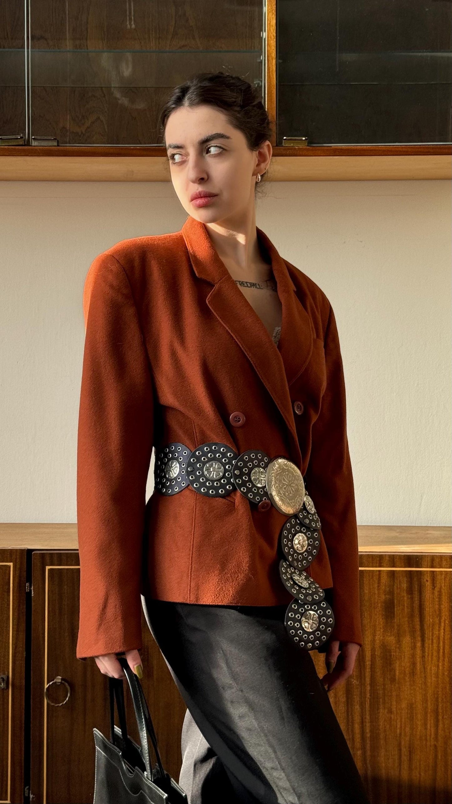 A woman wearing a brown blazer and carrying a black purse stands in front of a wooden cabinet.