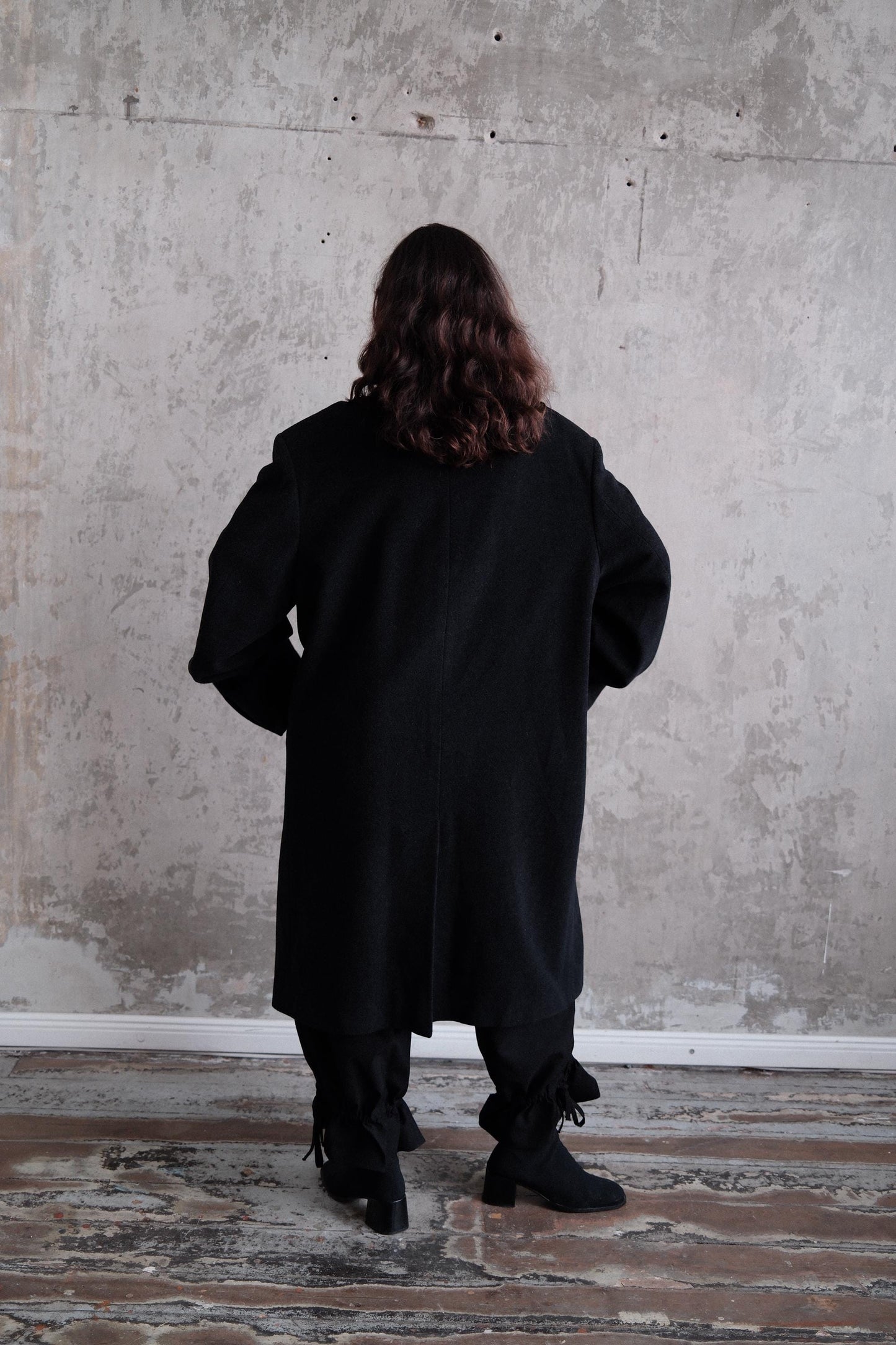 A person wearing a long black coat stands in a room with a concrete wall.