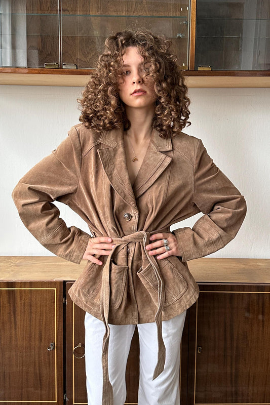 A woman with curly hair wearing a tan jacket stands confidently in front of a wooden cabinet.