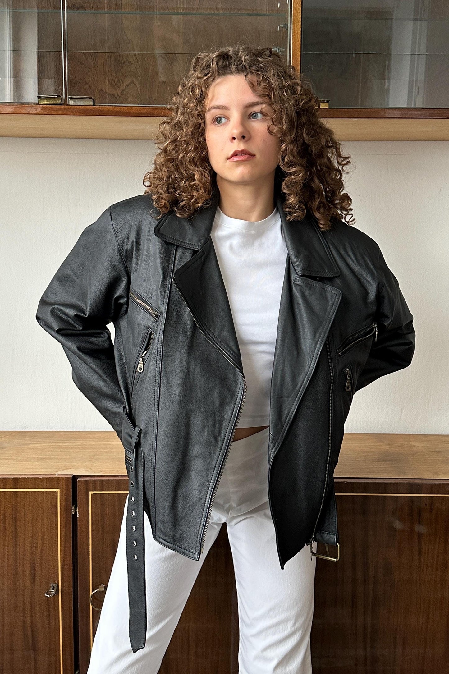 A young woman with curly hair wearing a black leather jacket stands in front of a wooden cabinet.