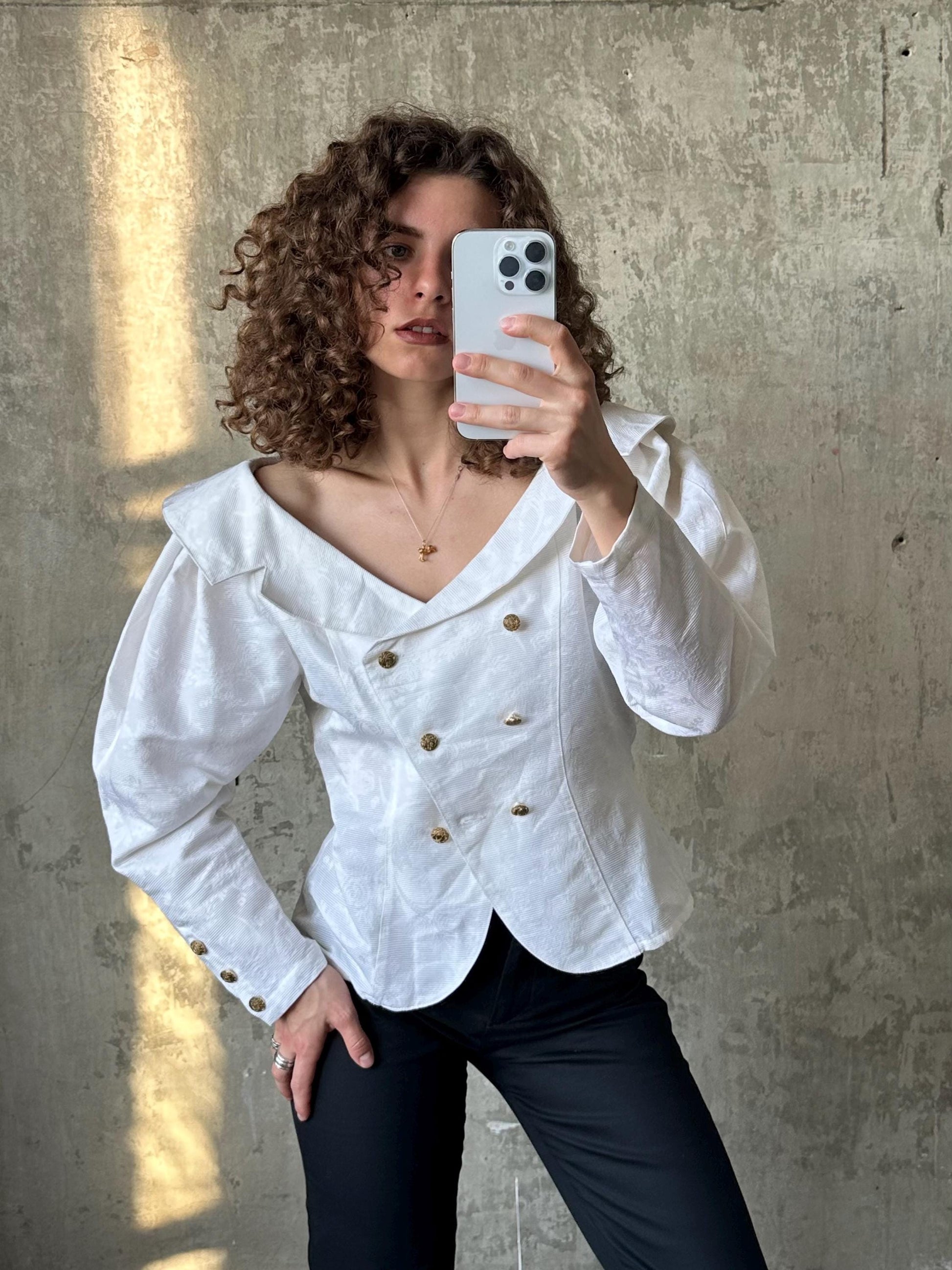 A woman with curly hair is taking a selfie with a white smartphone, wearing a white blouse with puffed sleeves and black pants.
