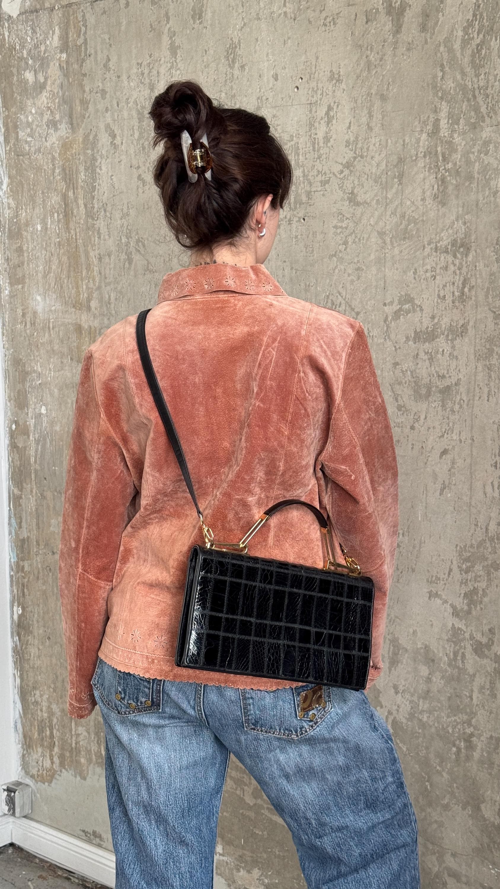 A woman wearing a pink jacket and blue jeans is standing against a concrete wall, carrying a black handbag with a crocodile skin texture.