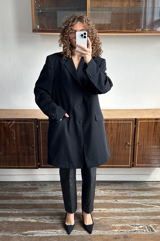 A person with curly hair is taking a selfie with a smartphone, wearing a black blazer and black pants.