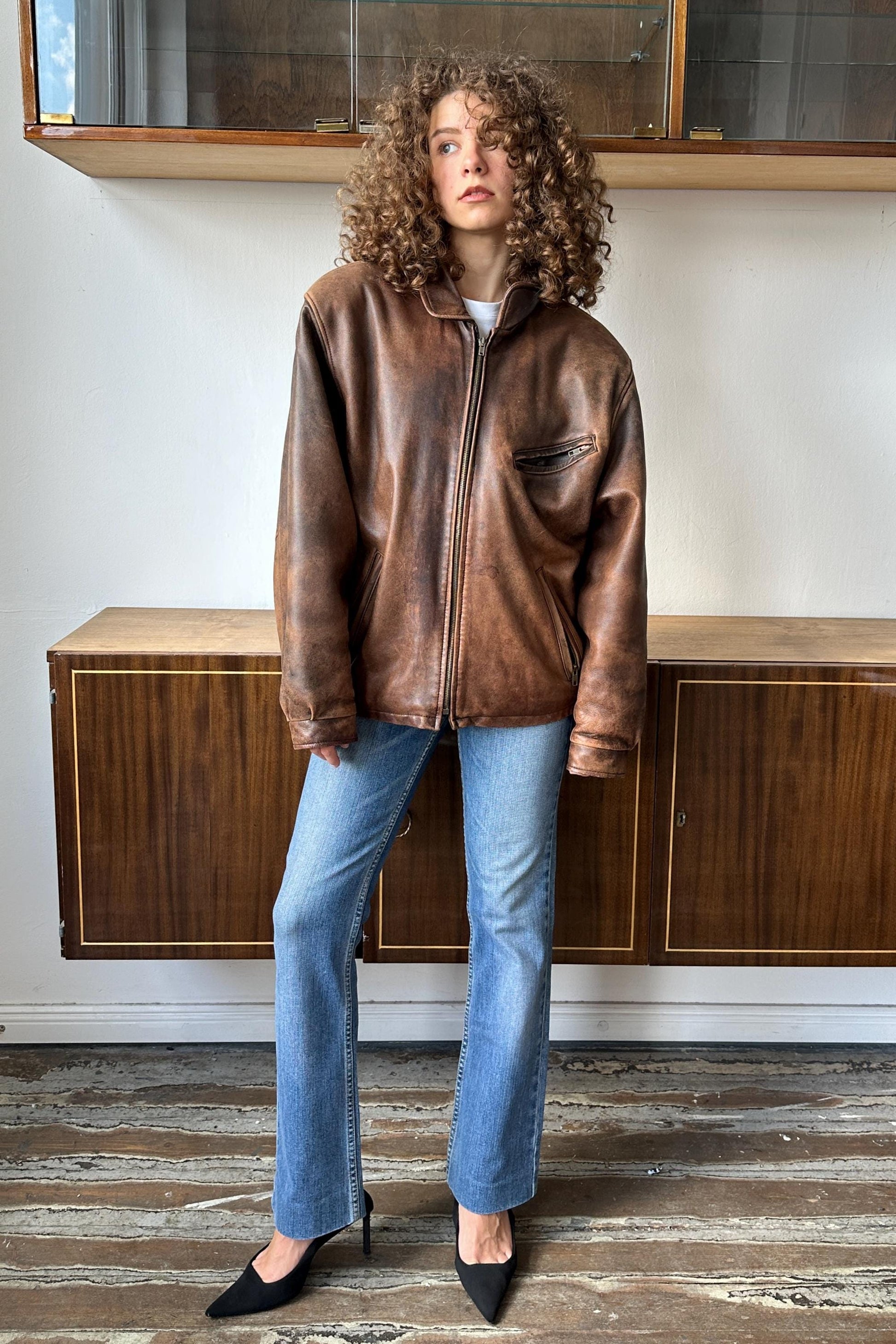 A woman with curly hair wearing a brown leather jacket and blue jeans stands in a room with wooden cabinets.