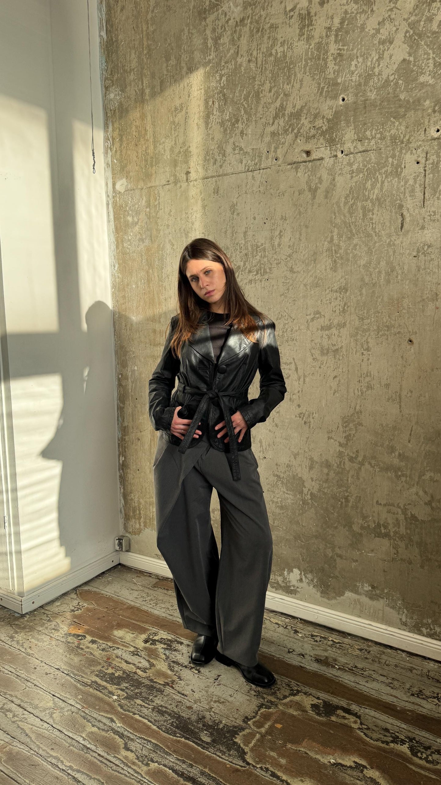 A woman in a black jacket and pants stands in a room with a concrete wall and wooden floor.