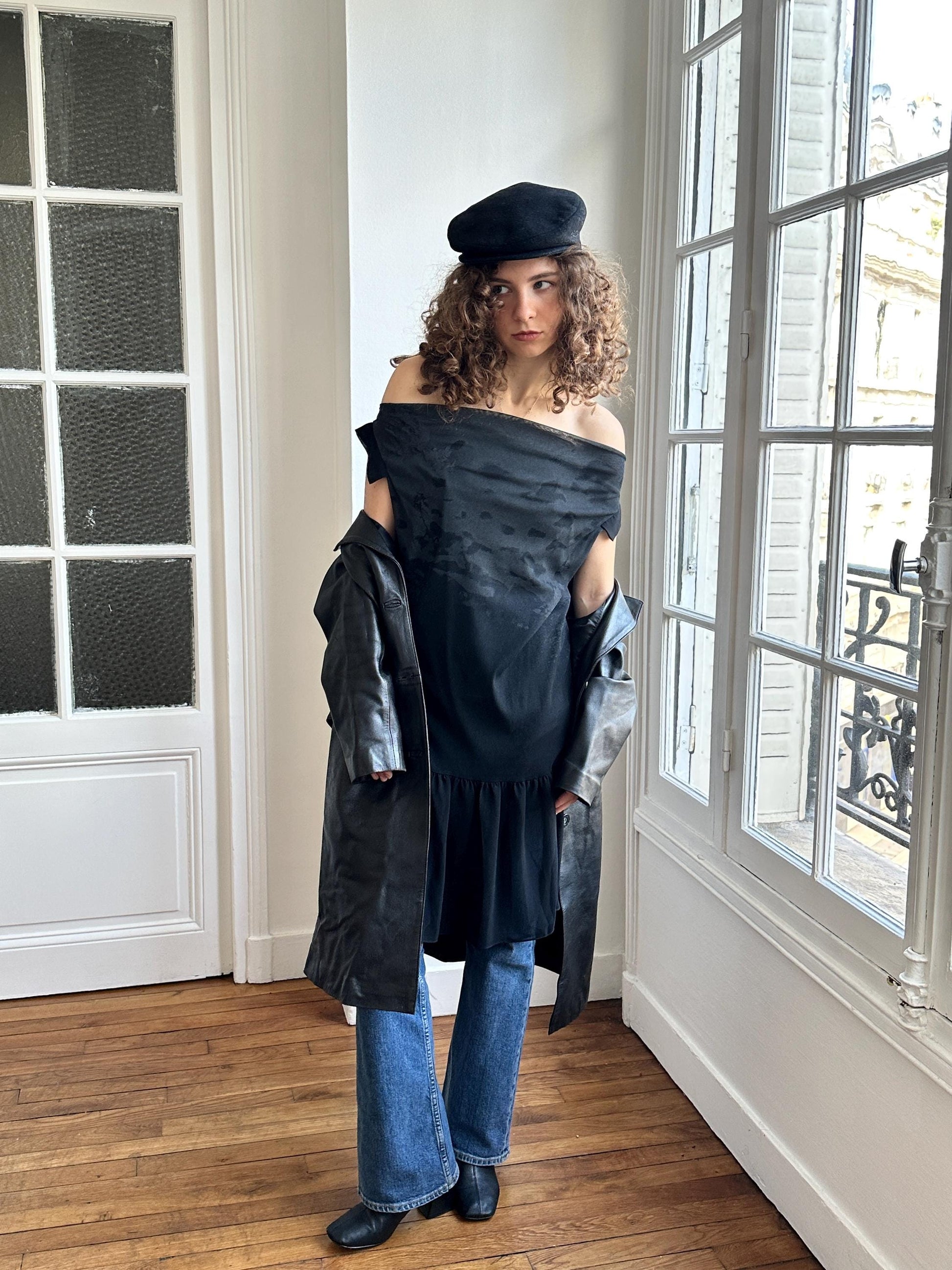 A woman wearing a black off-the-shoulder dress, a black hat, and a black leather jacket stands in a room with a window.