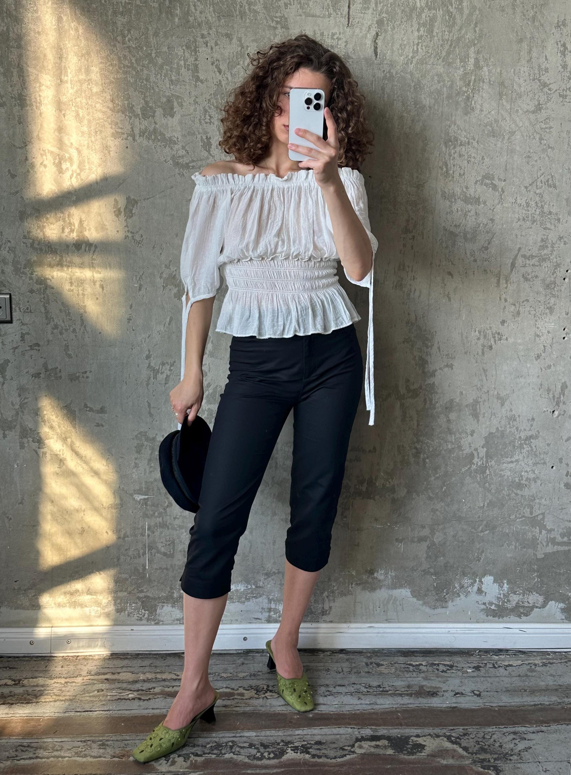 A woman with curly hair is taking a selfie with a white phone, standing in front of a concrete wall. She is wearing a white off-the-shoulder top, black pants, and green shoes.