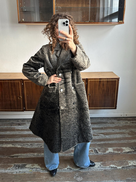 A person is taking a selfie with a smartphone, wearing a gray coat and blue jeans, standing in front of a wooden cabinet.