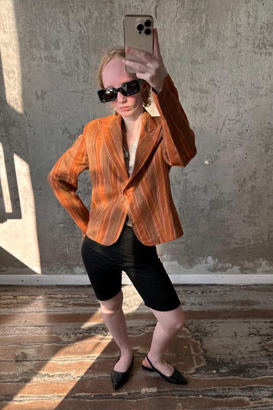 A person wearing a striped orange jacket, sunglasses, and black shorts is taking a selfie with a smartphone.