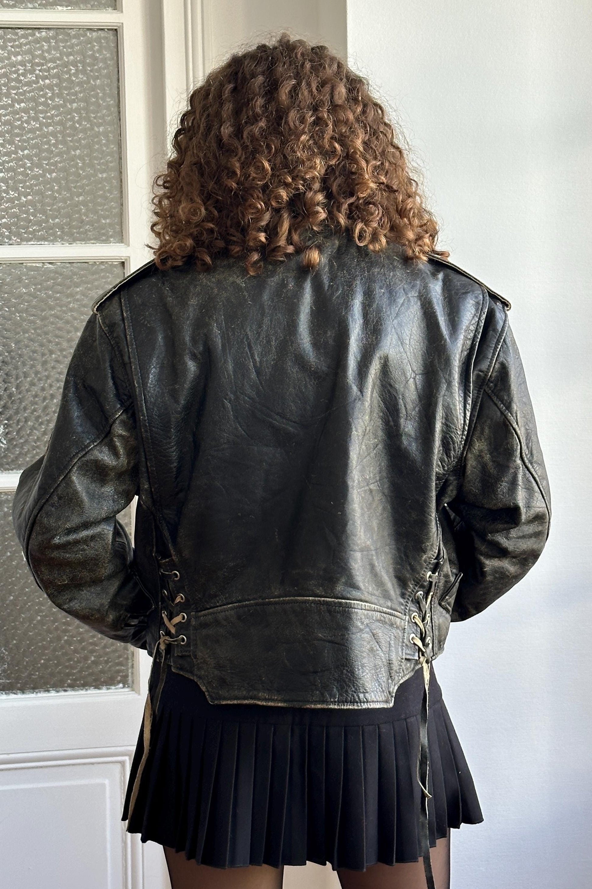 a person wearing a black leather jacket and a black skirt, standing in front of a window.