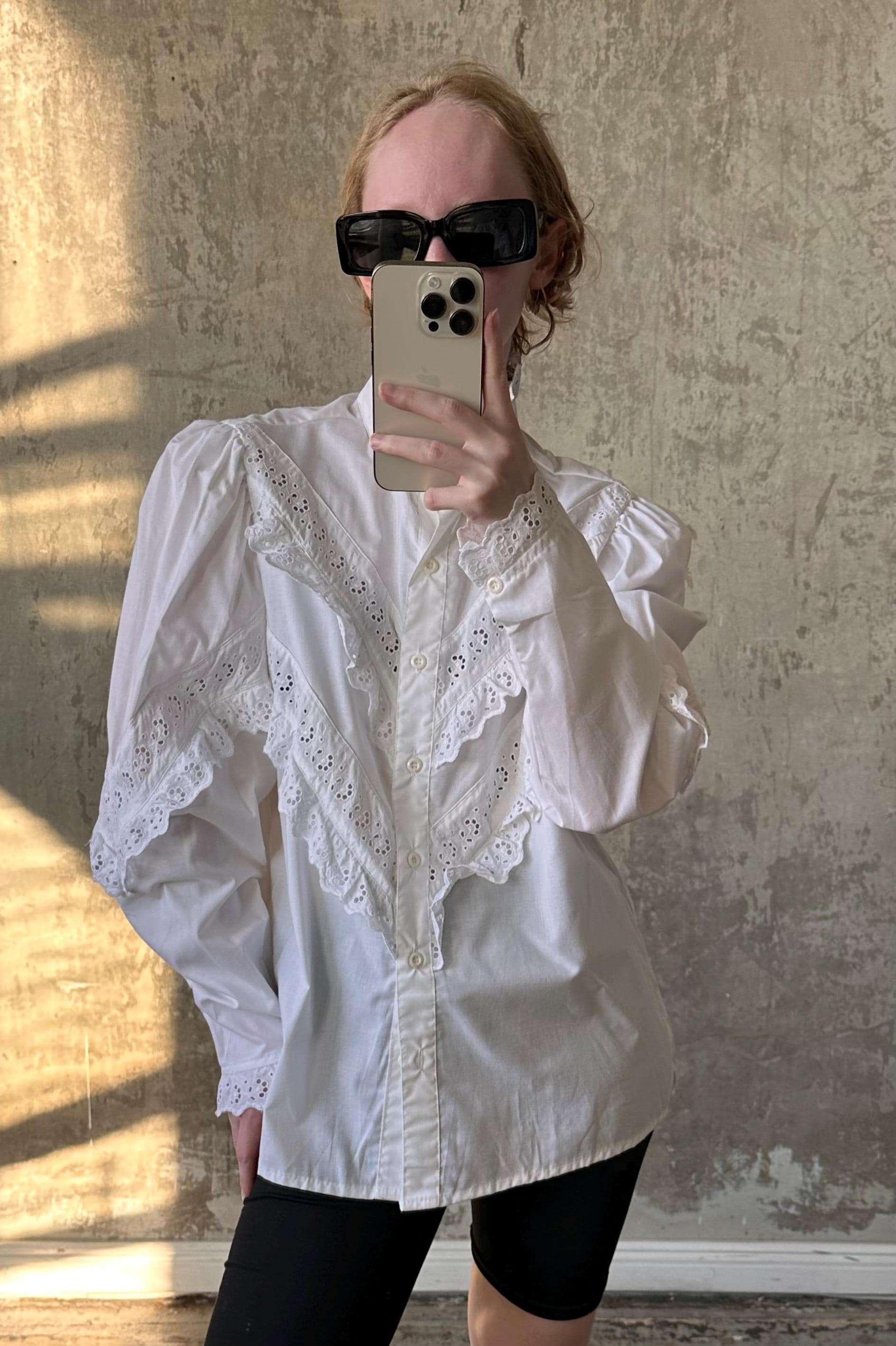 A person is taking a selfie with a smartphone, wearing a white lace blouse and sunglasses.
