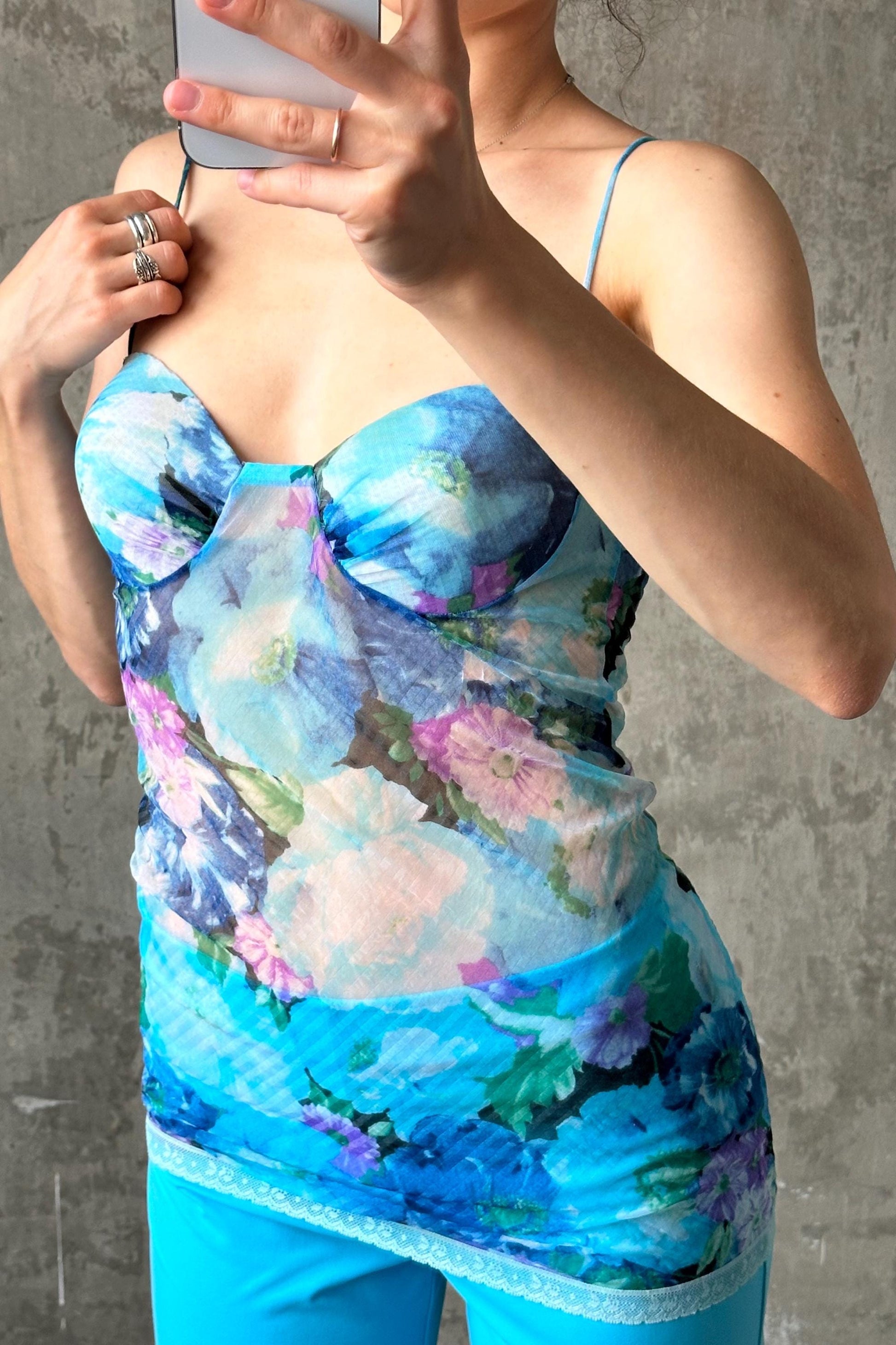 A woman is taking a selfie with a floral print top and blue pants, holding a smartphone in her hand.