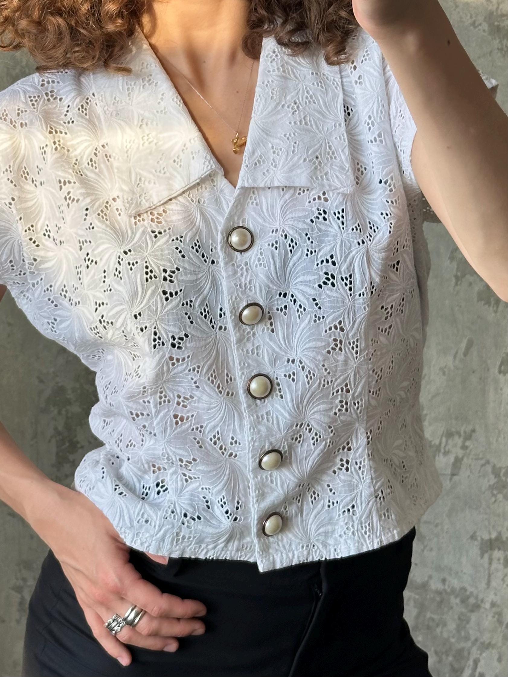 A close-up of a person wearing a white lace blouse with four buttons, posing with one hand on their hip.
