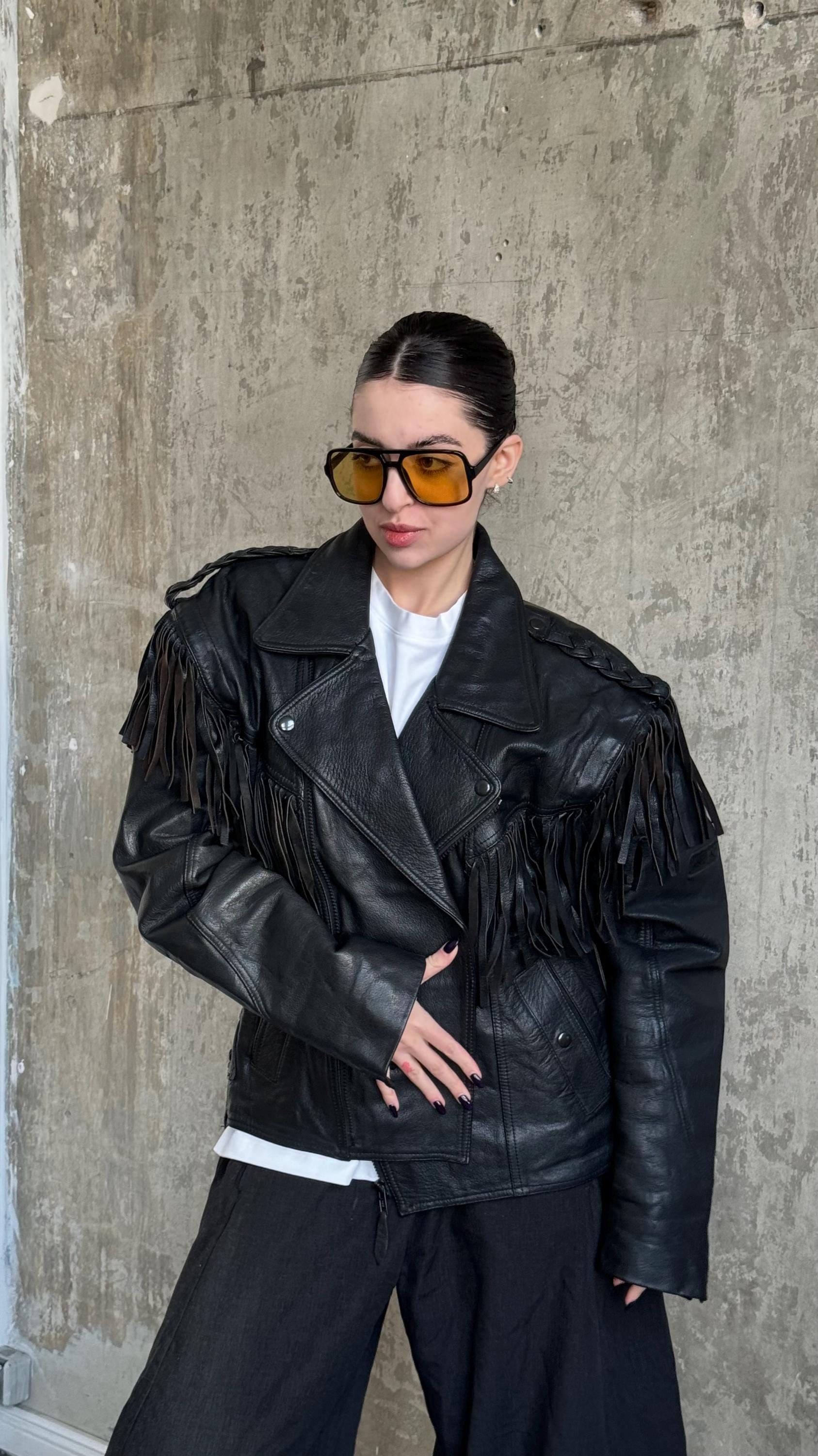 A woman wearing a black leather jacket with fringe, sunglasses, and standing in front of a concrete wall.