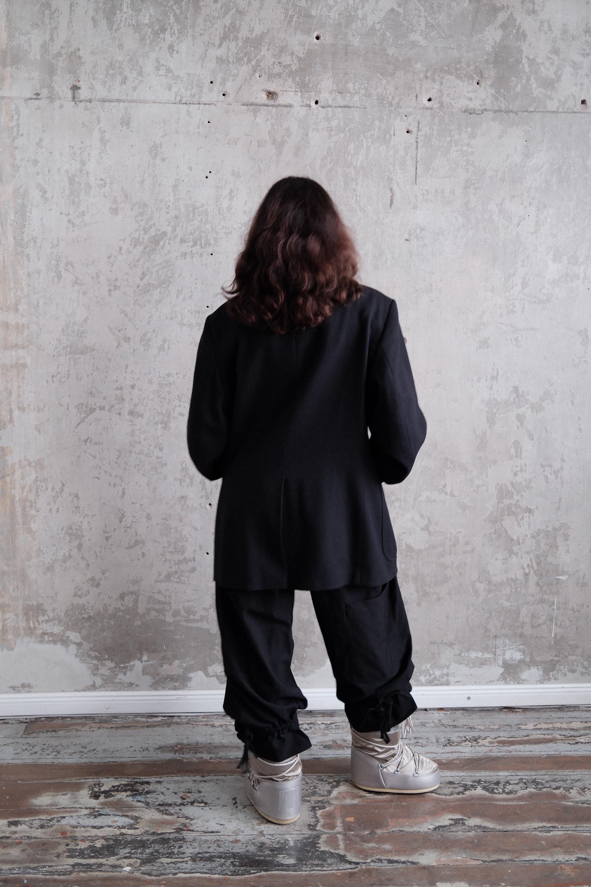a person standing in front of a concrete wall, wearing a black jacket and pants.