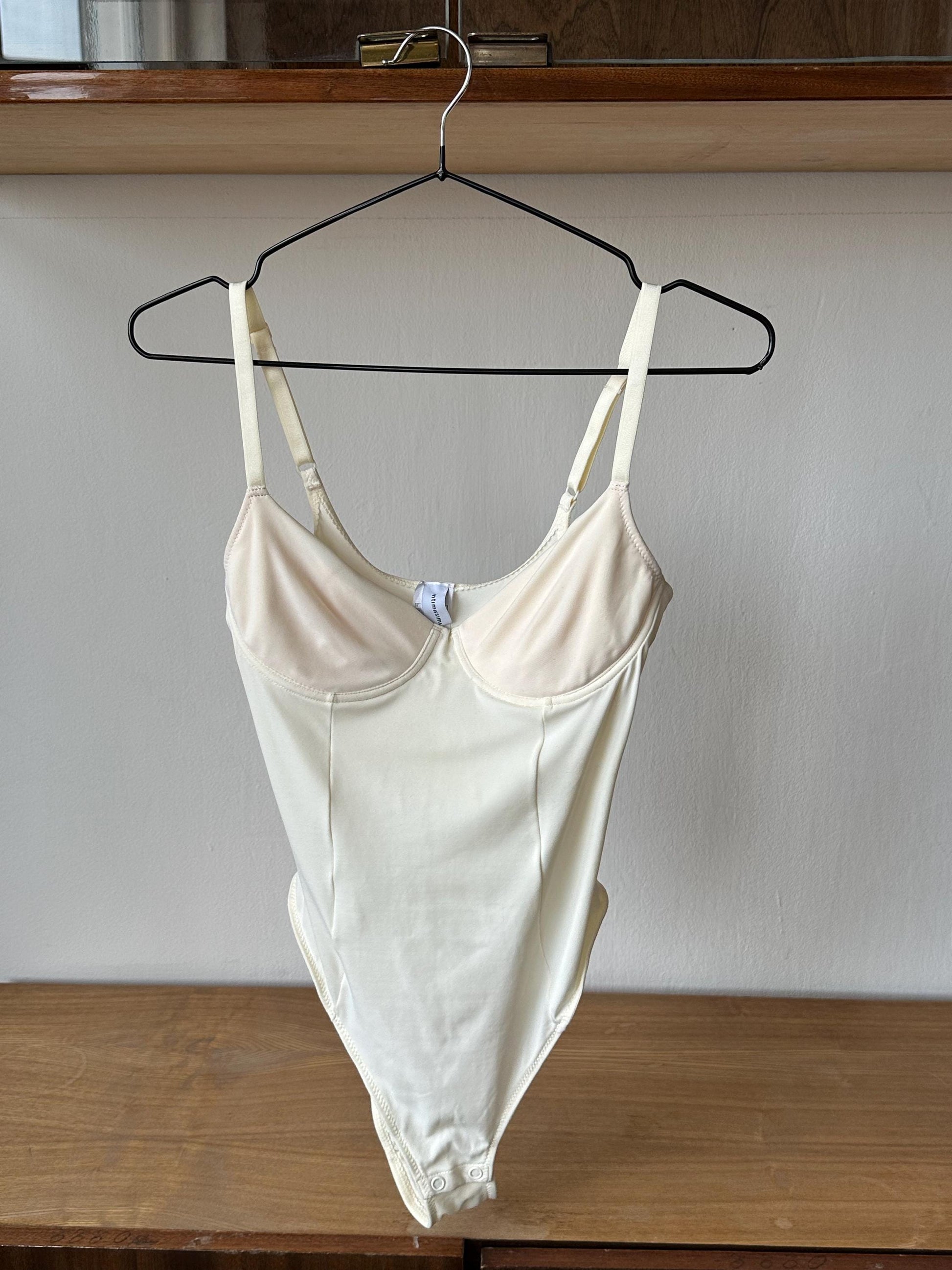 A white one-piece swimsuit is hanging on a black hanger on a wooden shelf.