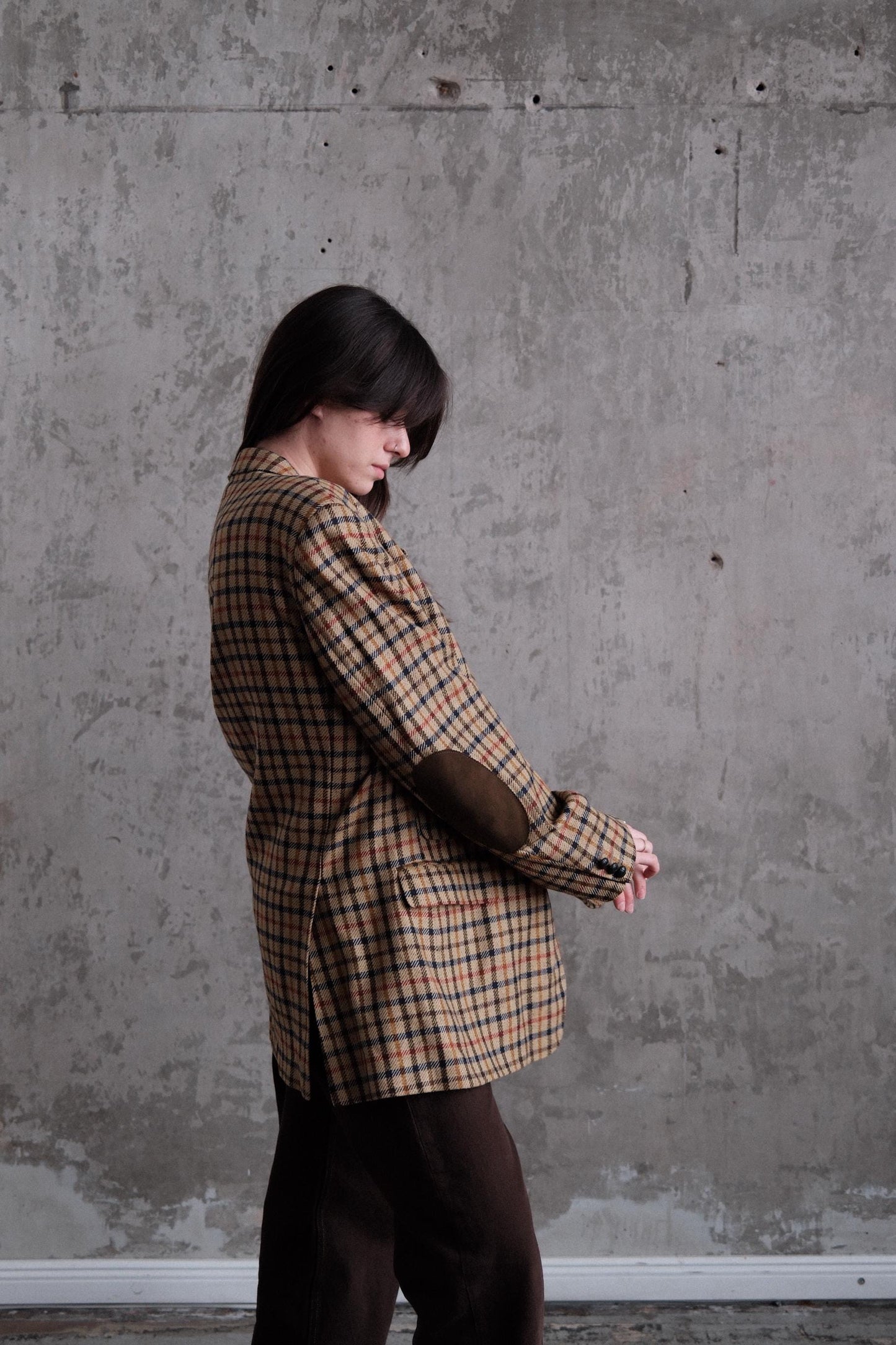 A person wearing a plaid jacket is bending over against a concrete wall.