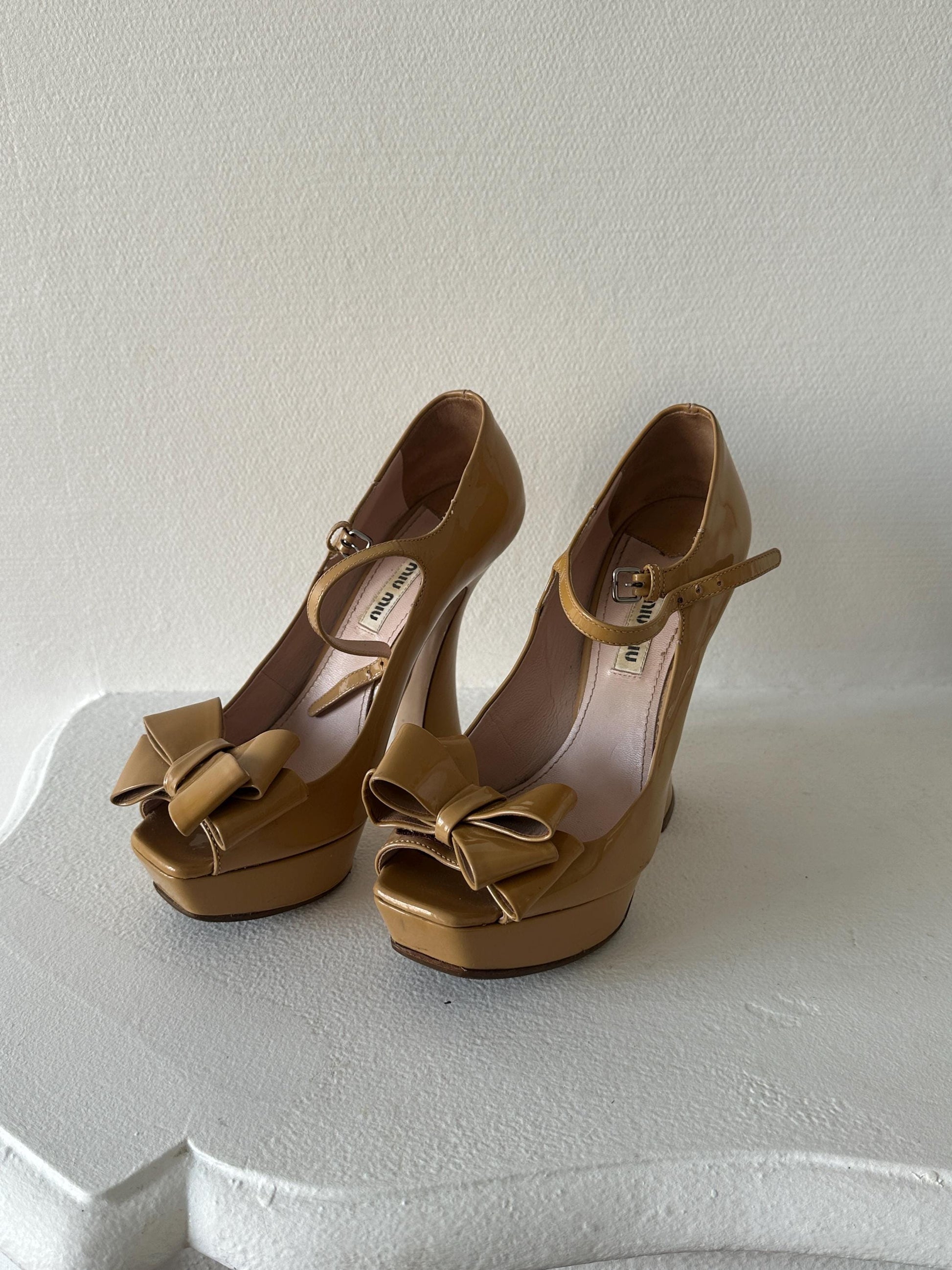 A pair of tan high-heeled shoes with bows on the toe and heel, placed on a white surface.