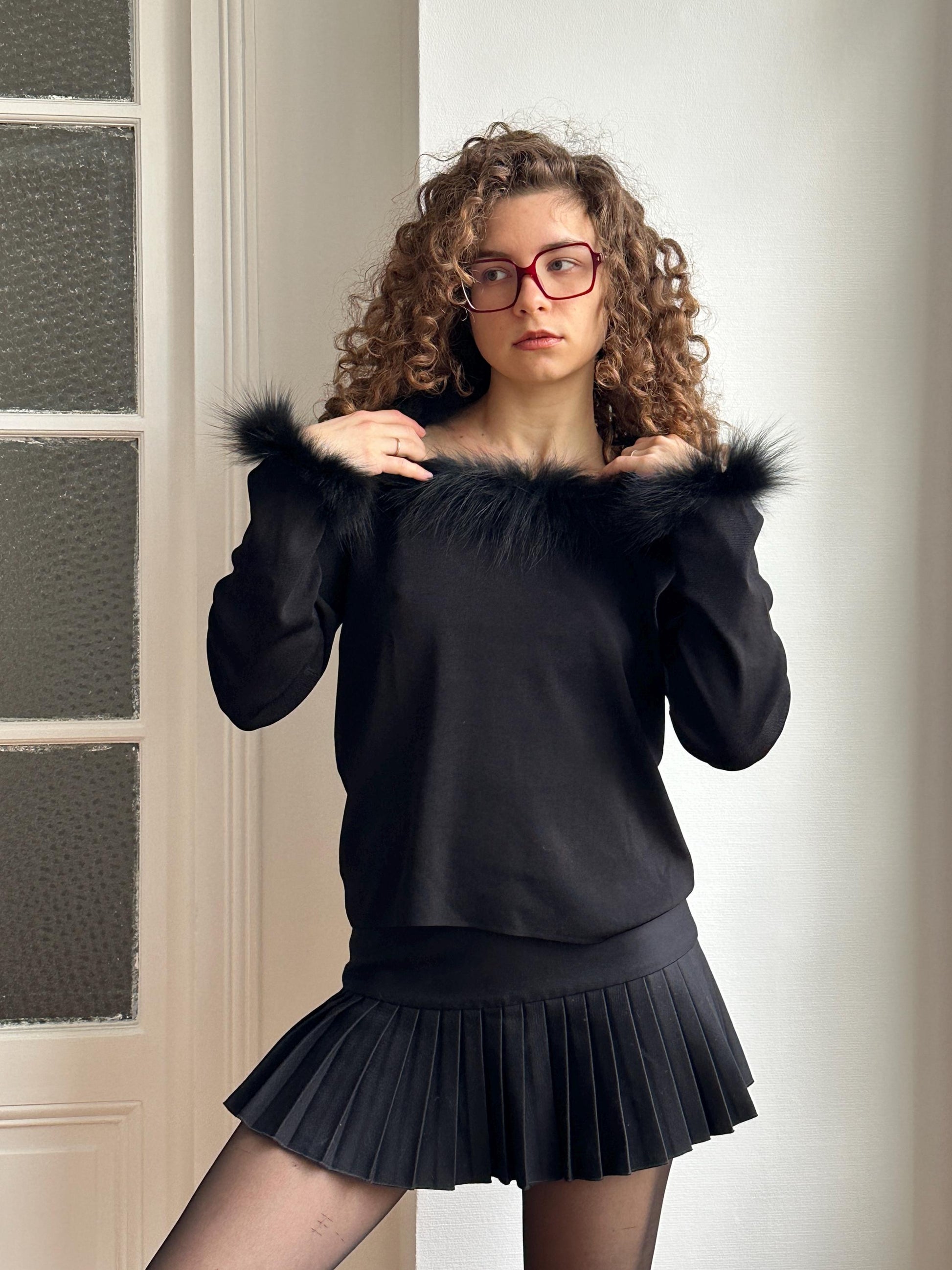 A woman with curly hair and glasses is wearing a black fur collar sweater and a black pleated skirt, posing in front of a white door.