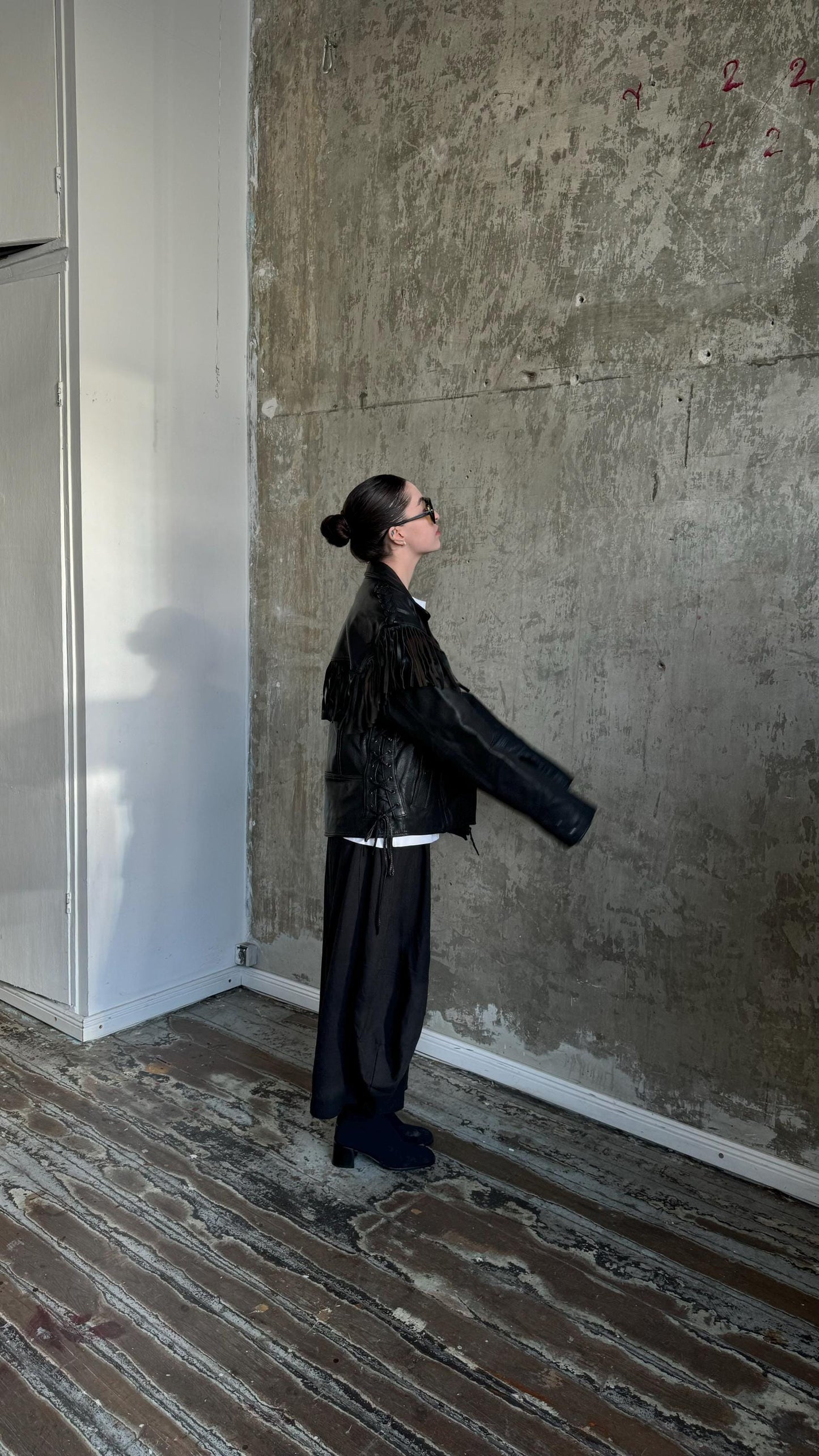 A person wearing a black leather jacket and sunglasses stands in a room with a concrete wall and wooden floor.