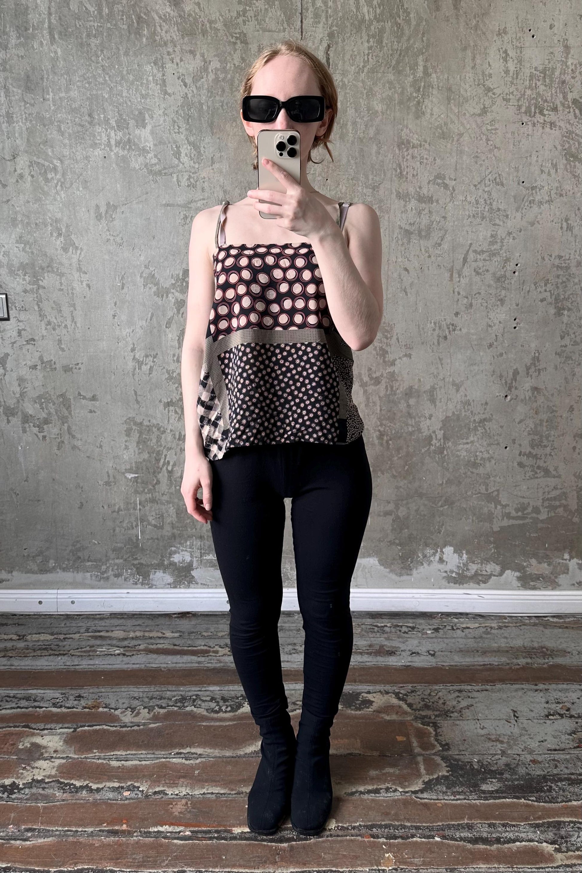 A woman wearing sunglasses and a patterned top is taking a selfie with a smartphone in front of a concrete wall.