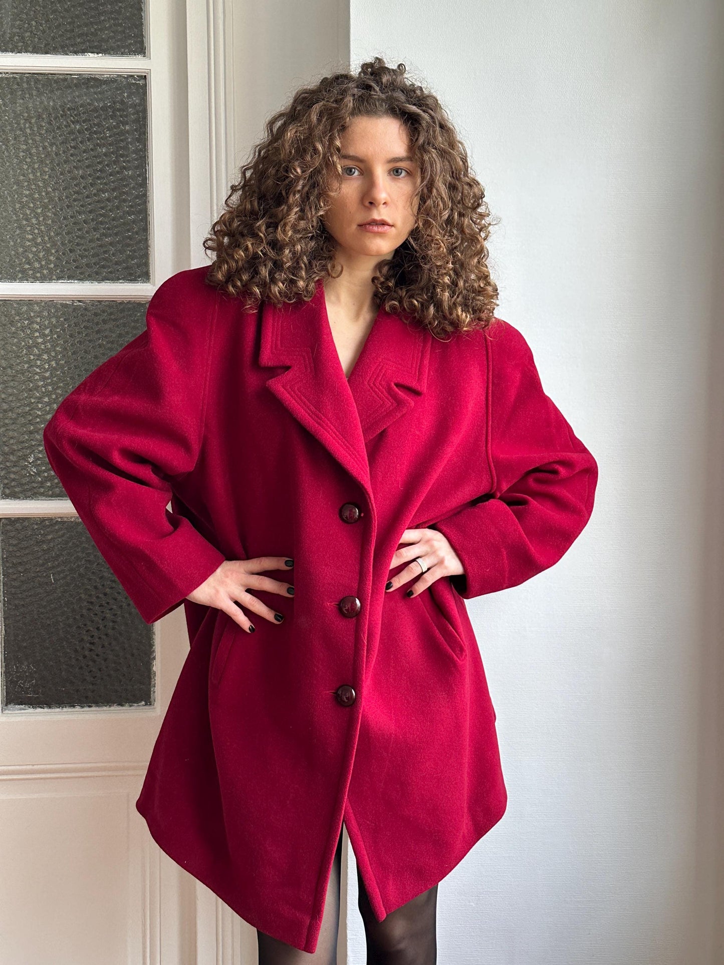 A woman with curly hair wearing a red coat is posing in a relaxed manner.