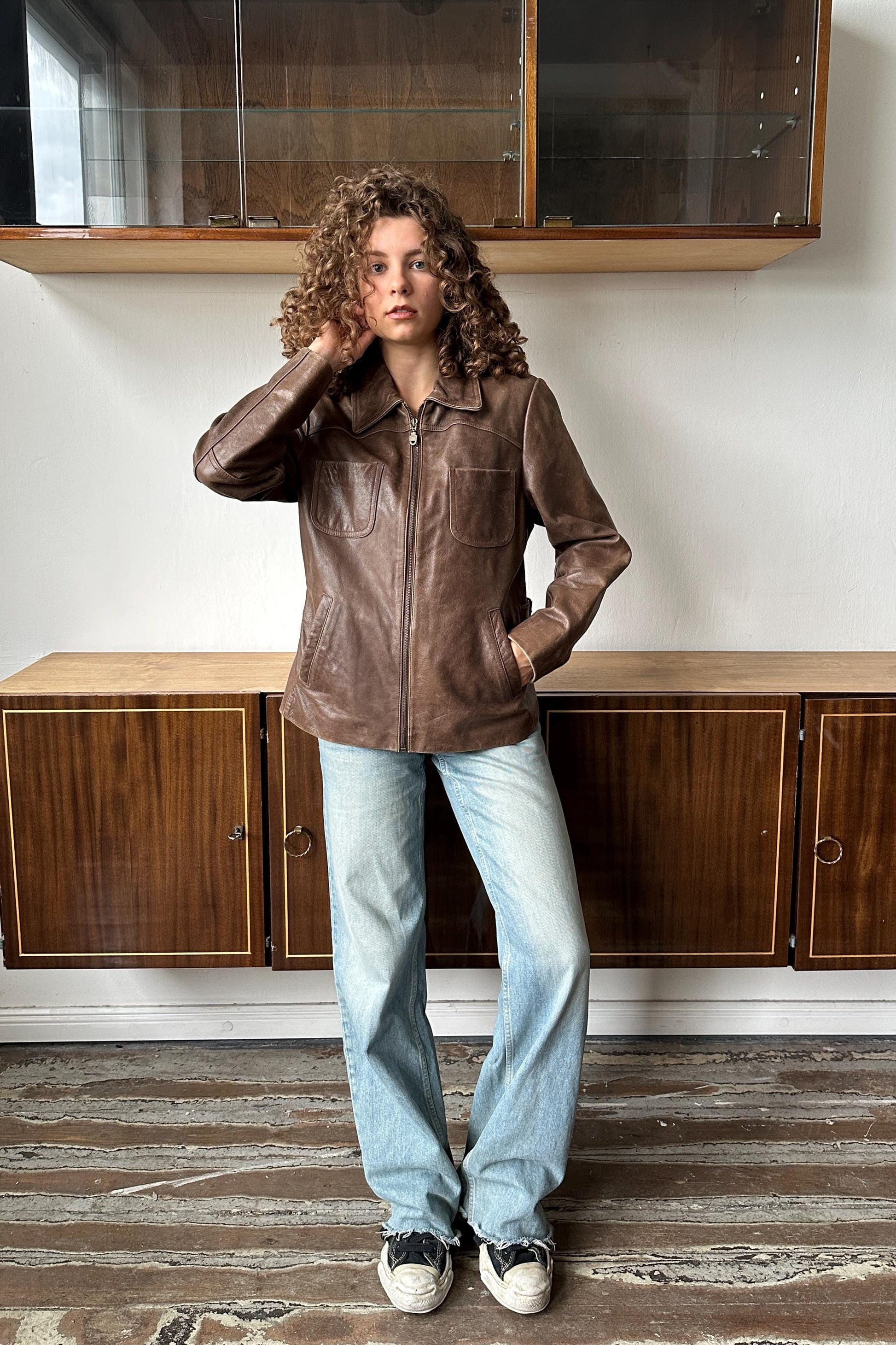 A woman with curly hair stands in a room, wearing a brown leather jacket and blue jeans.