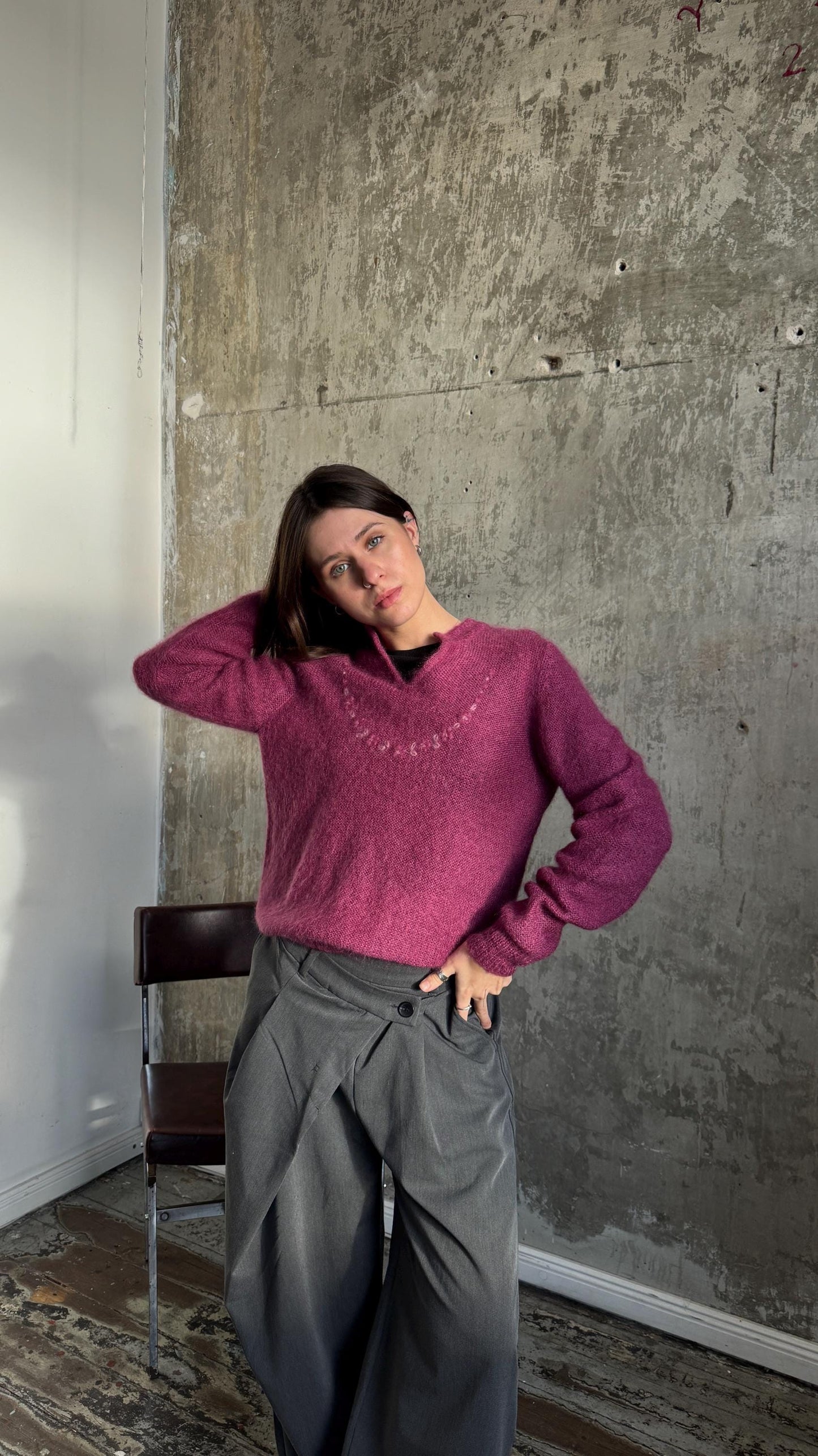 A woman wearing a pink sweater and gray pants stands in front of a concrete wall, with her hand on her head.