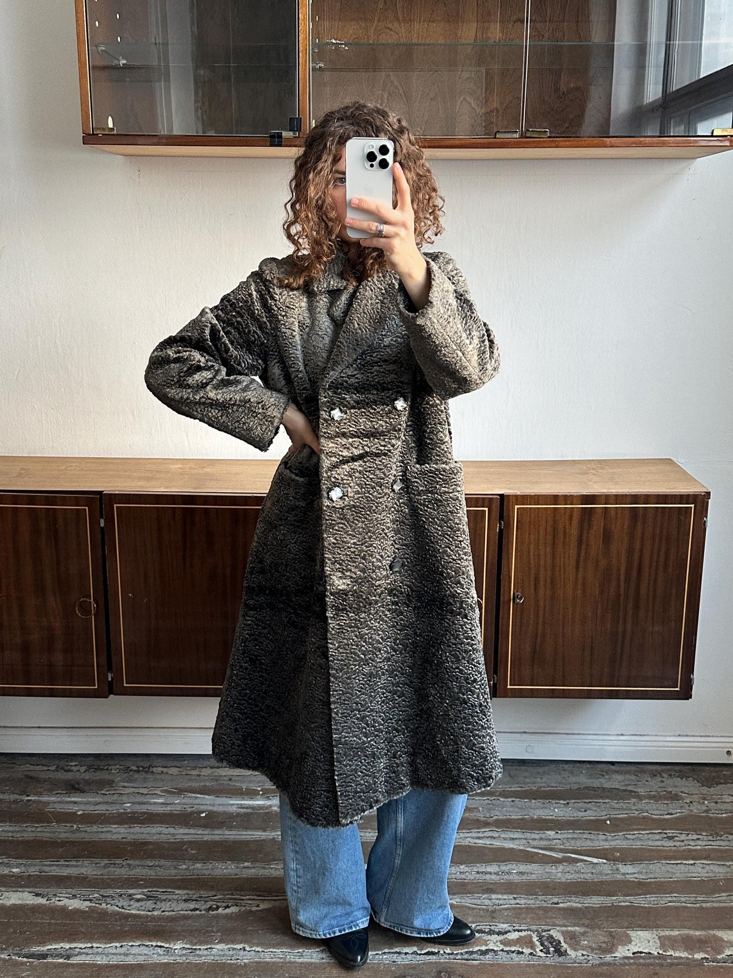 A person is taking a selfie with a smartphone, wearing a gray coat and blue jeans, standing in front of a wooden cabinet.