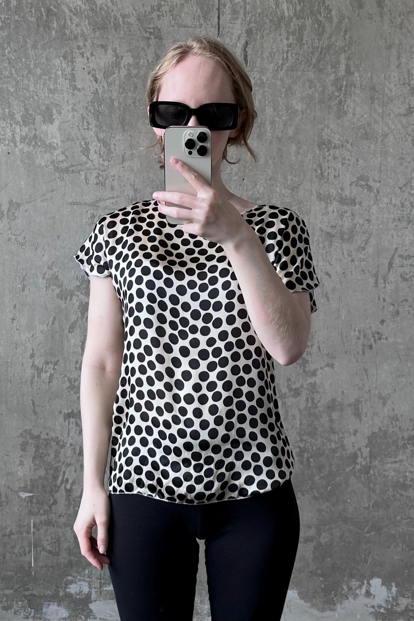 A woman wearing a black and white polka dot blouse and sunglasses is taking a selfie with her iPhone against a concrete wall.