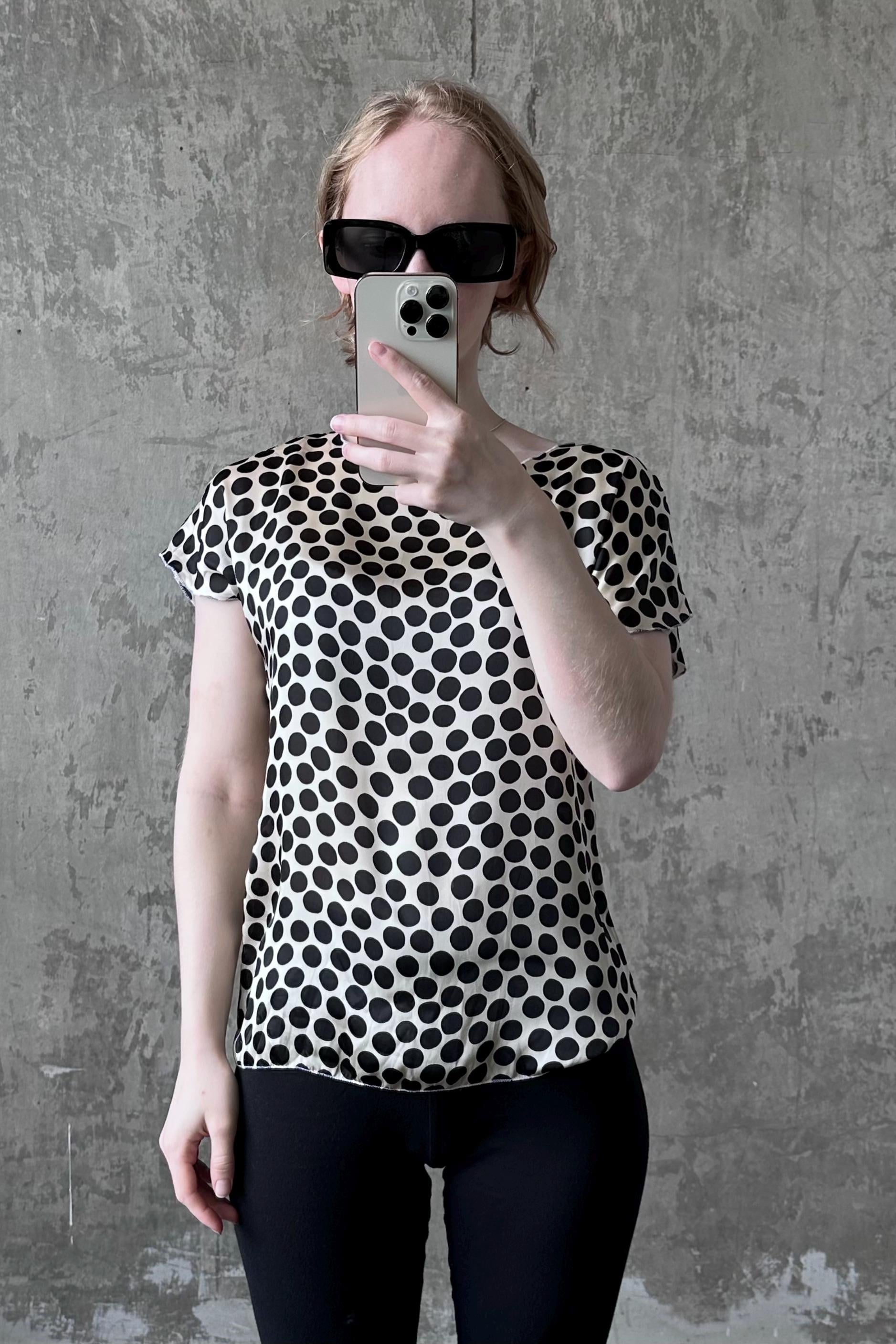 A woman wearing a black and white polka dot blouse and sunglasses is taking a selfie with her iPhone against a concrete wall.