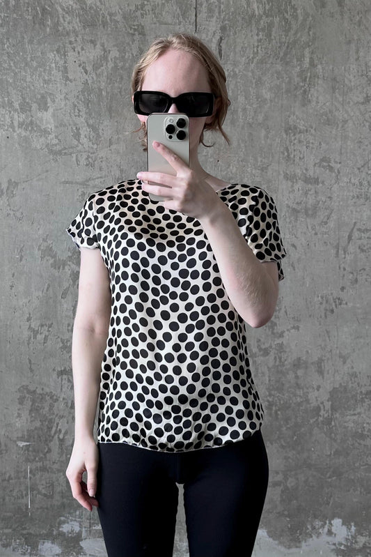 A woman wearing a black and white polka dot blouse and sunglasses is taking a selfie with her iPhone against a concrete wall.