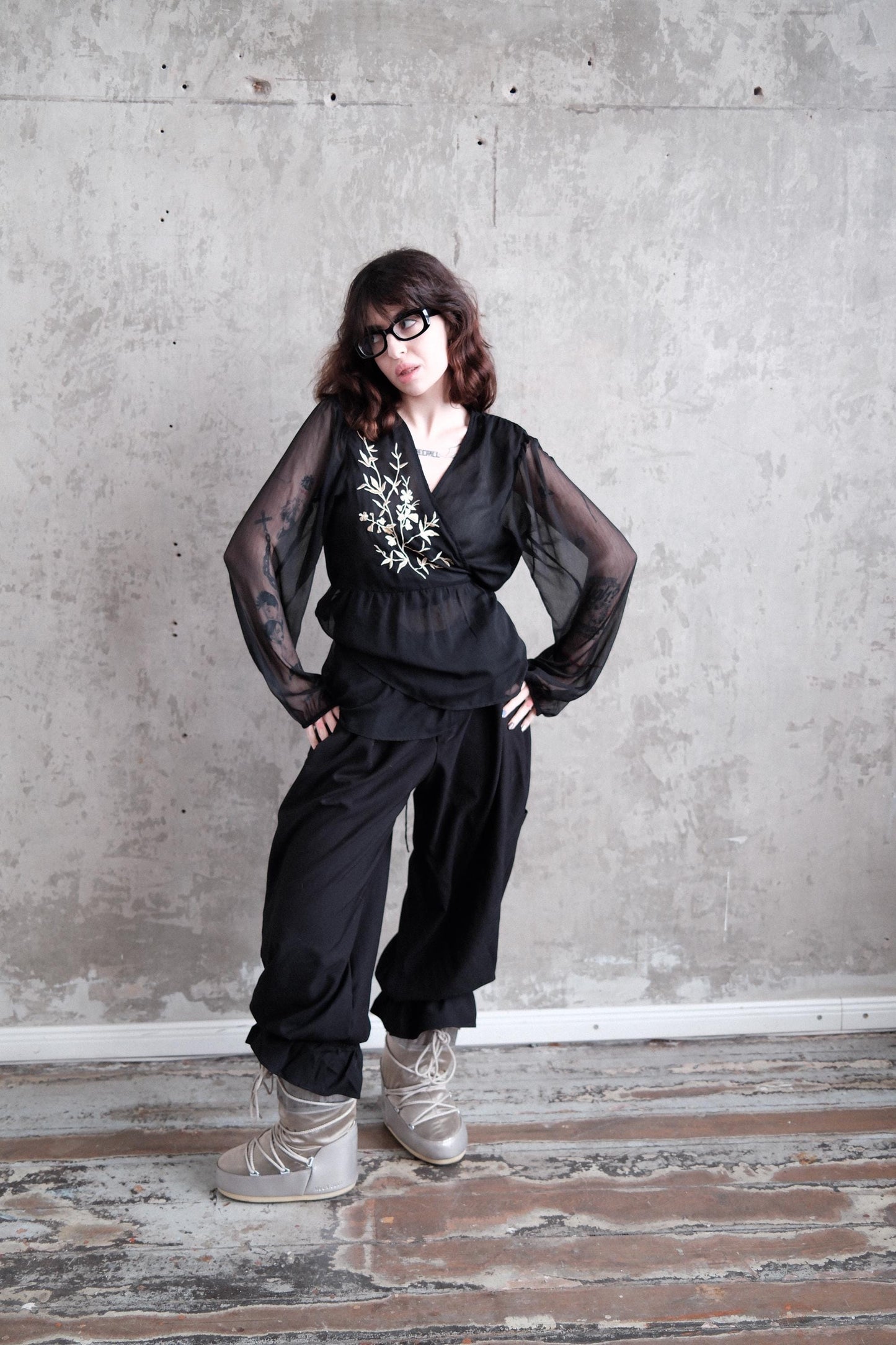 A woman in a black outfit with sheer sleeves stands confidently with her hands on her hips, wearing glasses and posing against a plain concrete wall.