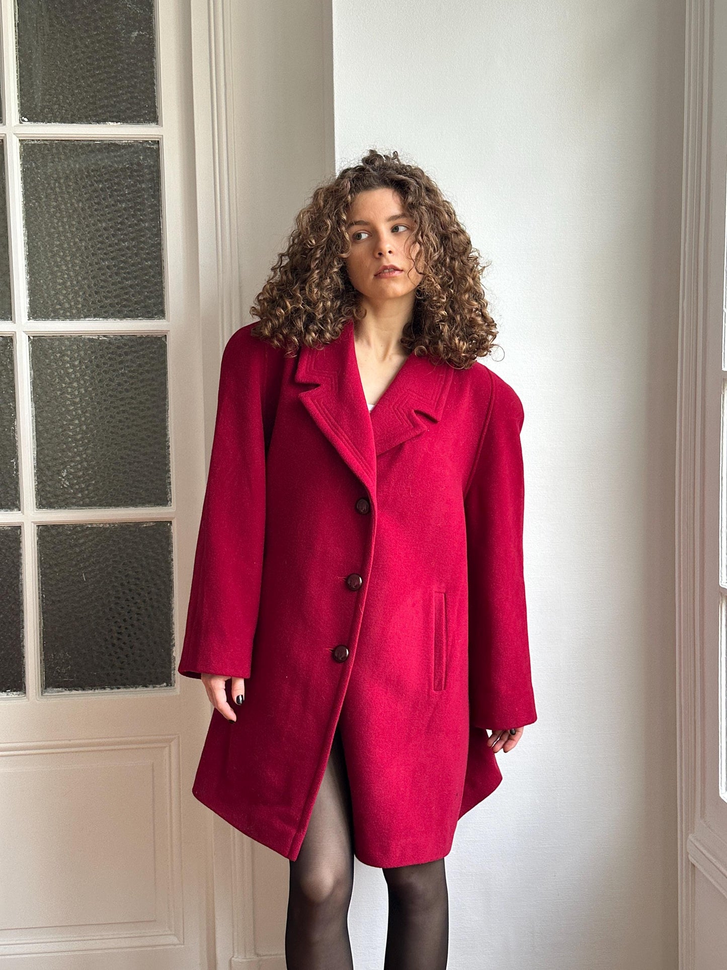 A woman with curly hair wearing a red coat stands in front of a white wall with a window.