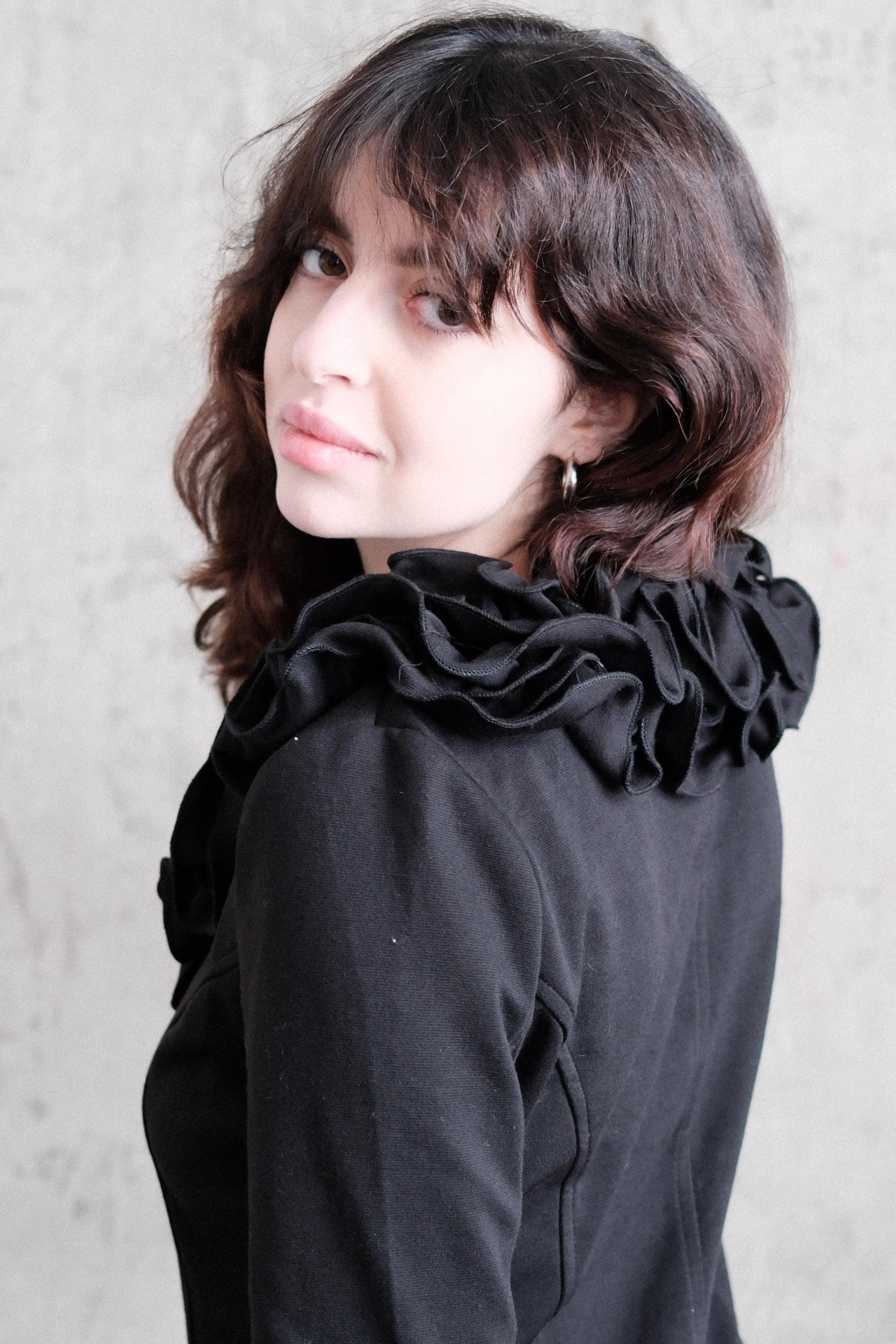 A young woman with long, wavy brown hair wearing a black ruffled jacket is looking up at the camera.