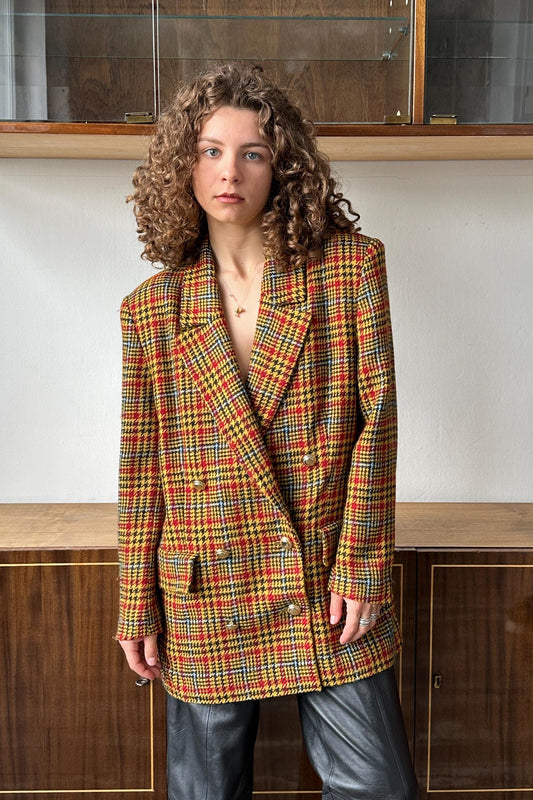 A woman with curly hair wearing a colorful, patterned jacket stands in front of a wooden cabinet.