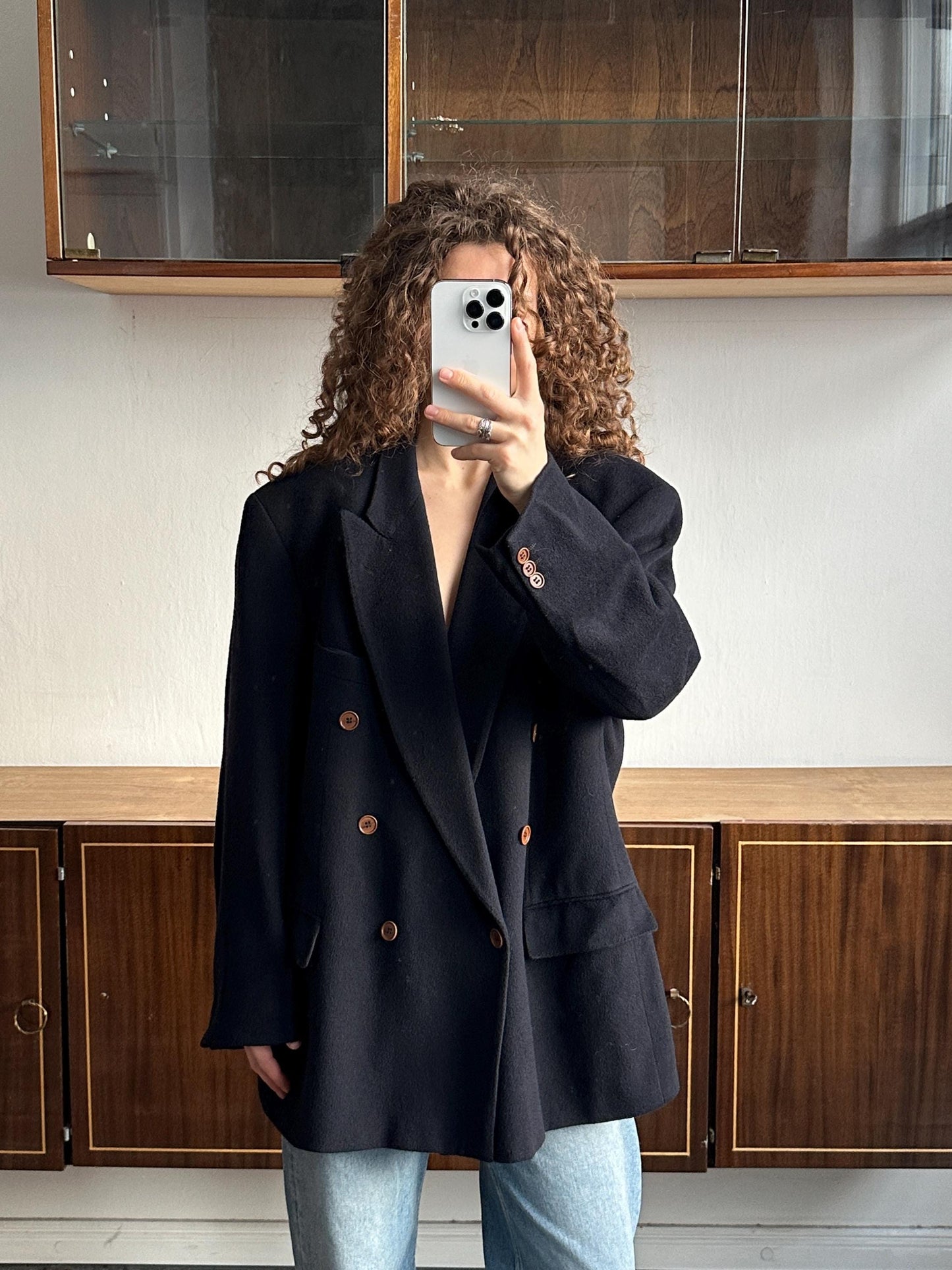 A person with curly hair is taking a selfie with a white phone, wearing a black blazer and blue jeans.