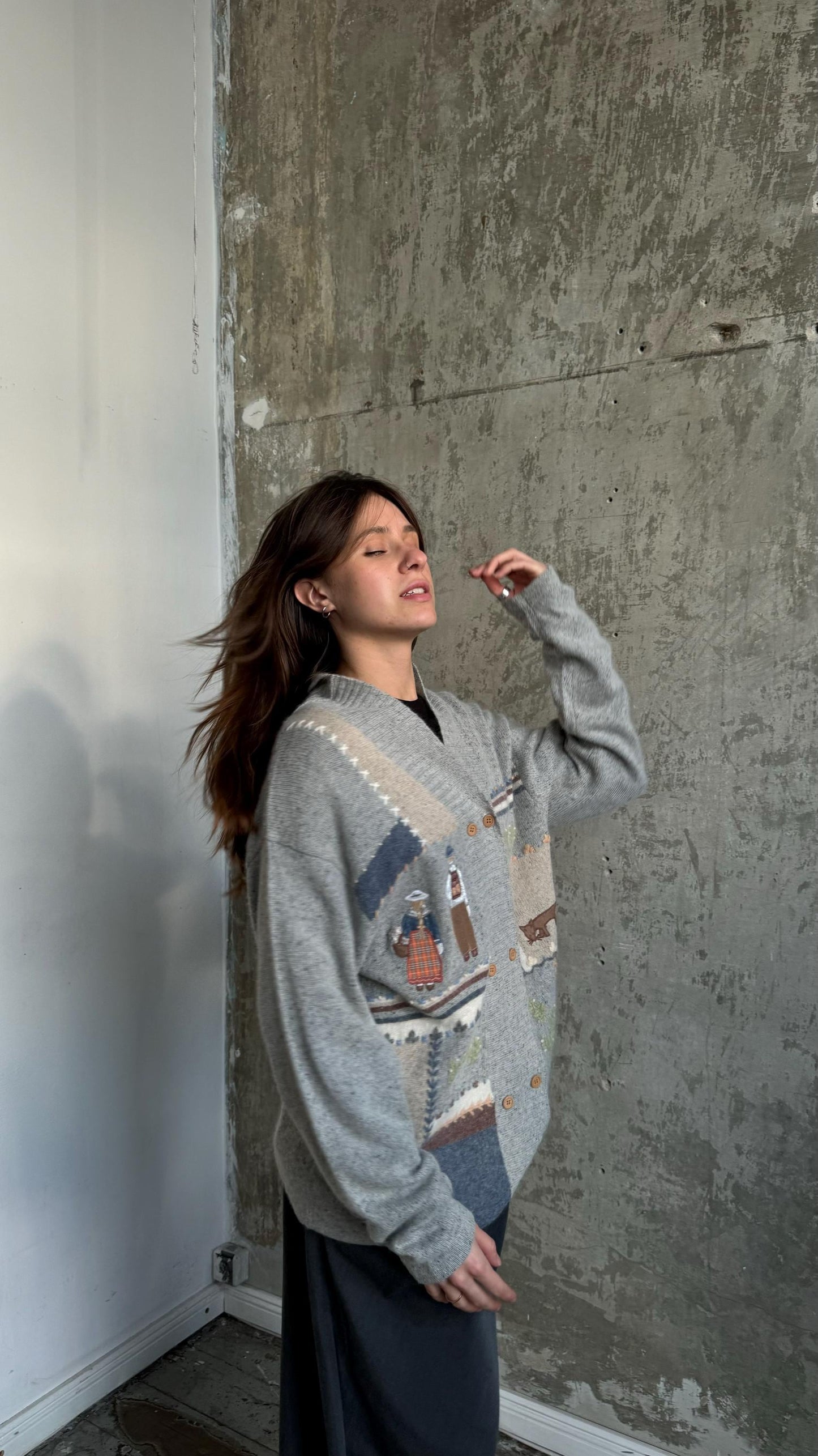 A woman wearing a gray sweater with a patterned design stands in front of a concrete wall, her hand touching her face.