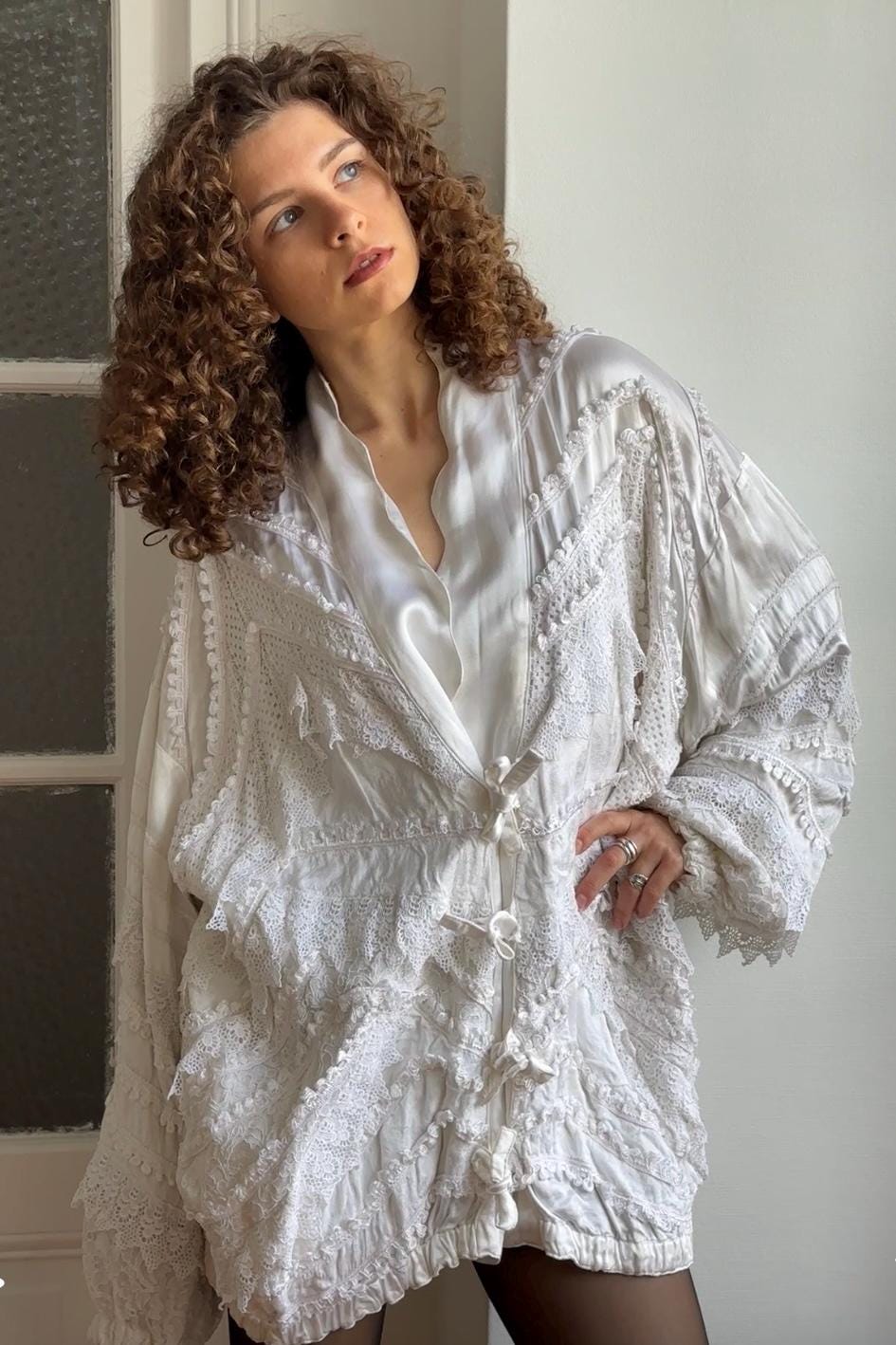 A woman with curly hair wearing a white, ruffled jacket stands in front of a window.