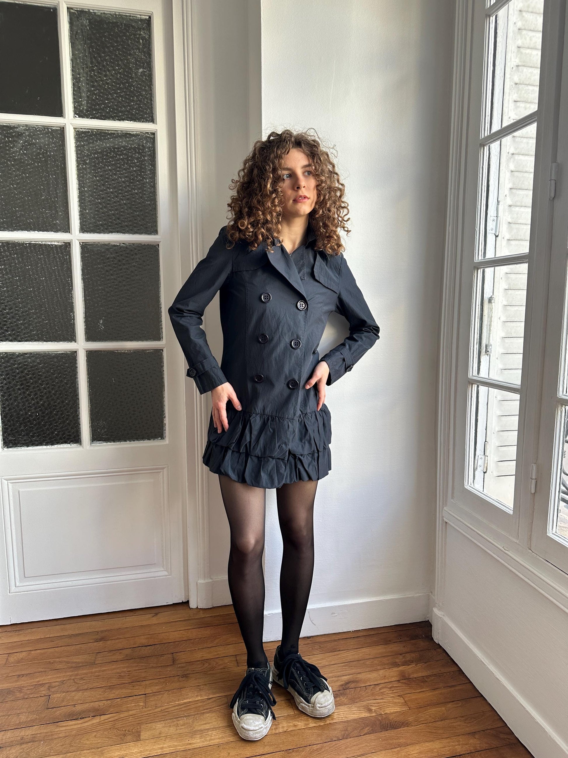 A woman with curly hair stands in a room with a window, wearing a black coat and black shoes.