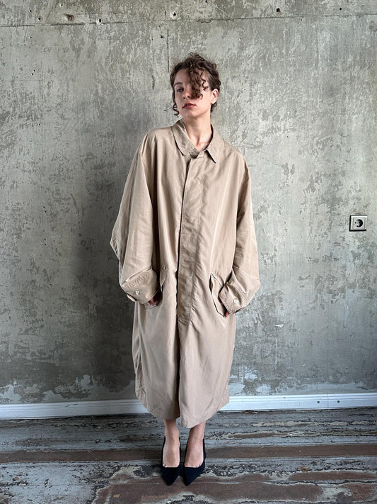 A woman wearing a beige trench coat stands in front of a concrete wall, looking off to the side with a neutral expression.
